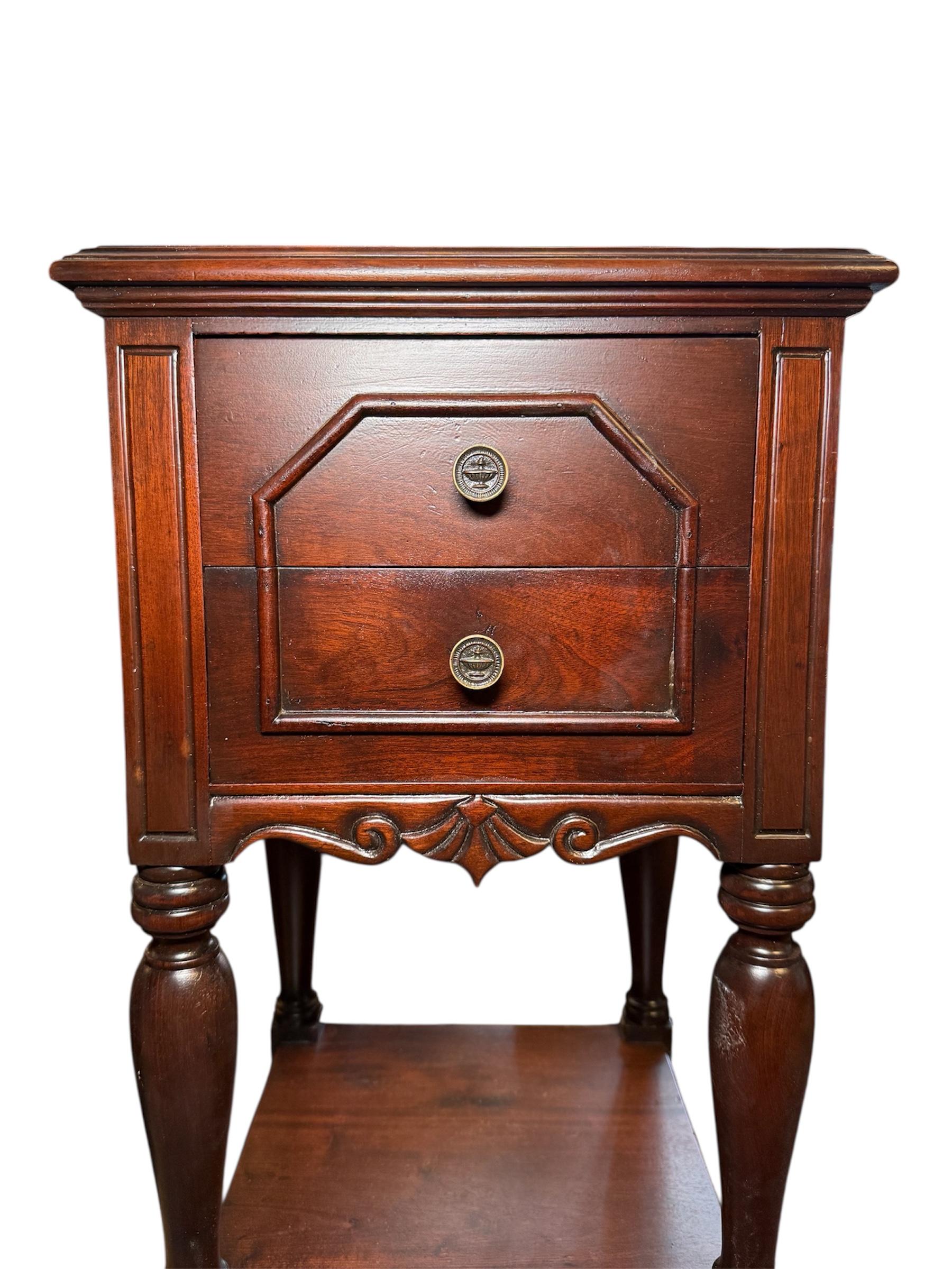 This is an American Colonial Style Wood Bedside Table/Nightstand. It depicts a dark brown hand painted wood square top shaped small table. It is supported by four balusters legs that also hold a lower shelf. The table consists of two drawers with a