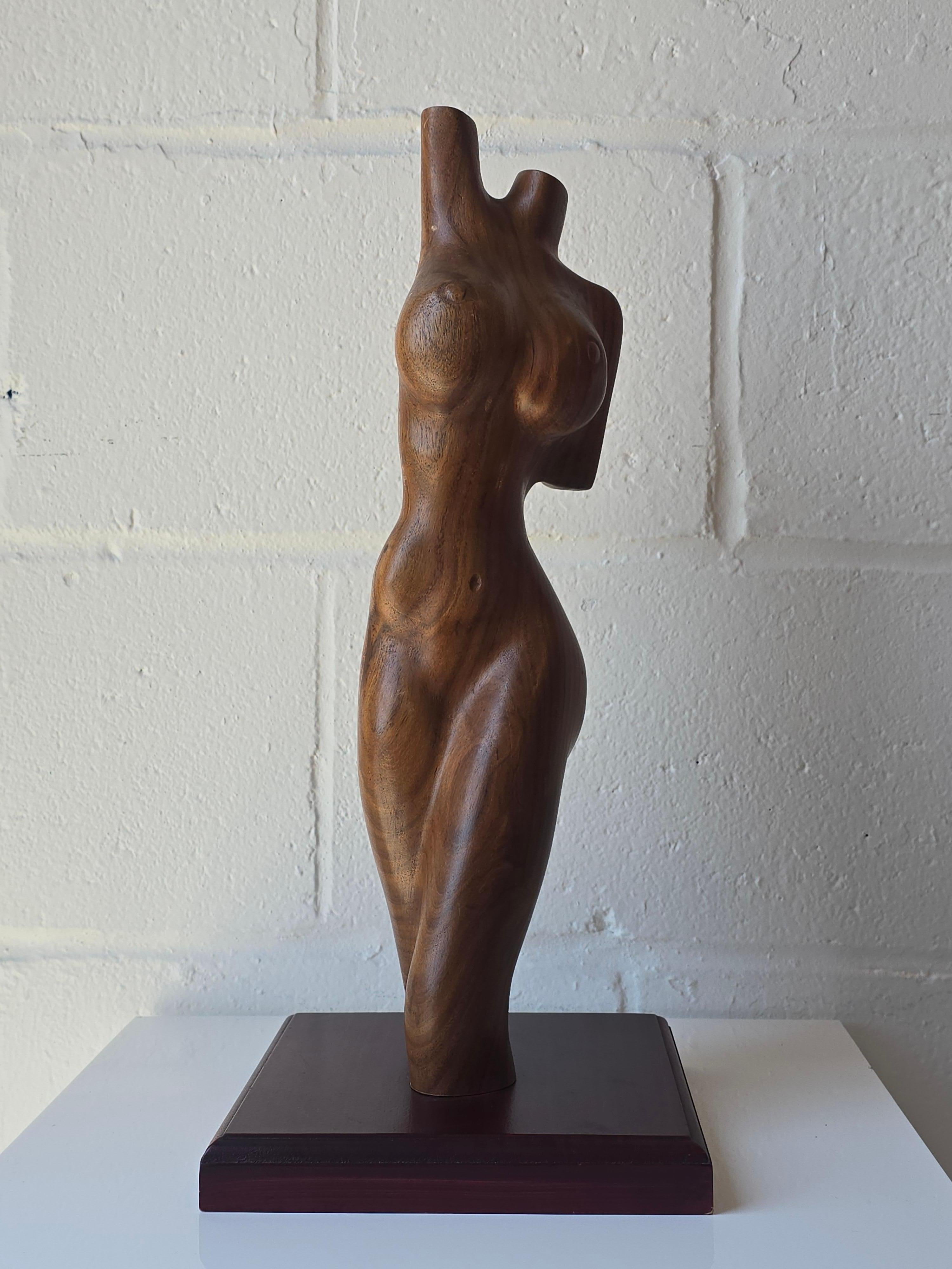 Una scultura di figura femminile in legno intagliata a mano. La scultura intagliata in legno ha la forma di una figura femminile e poggia su una base quadrata. Questa figura scultorea sembra essere realizzata in legno di noce, meticolosamente