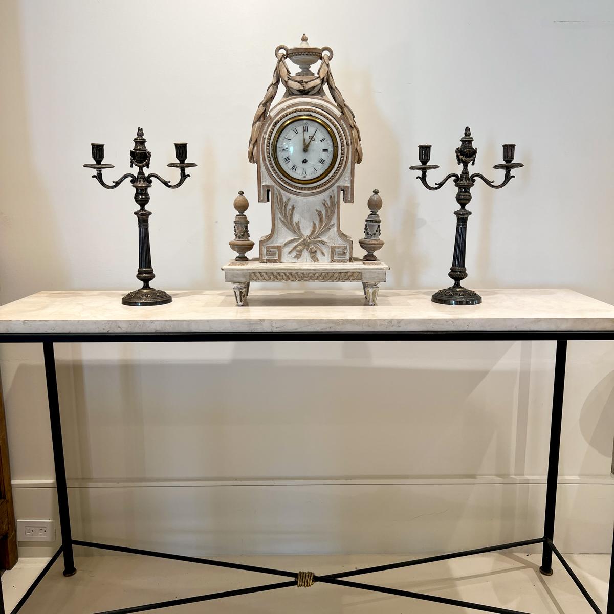 Very ornate white washed wood mantle clock with gold details. The face is behind a functional glass door with latch. The top is crowned by an urn with a cascading laurel garland that falls to either side of the case. There are pretty finials at the