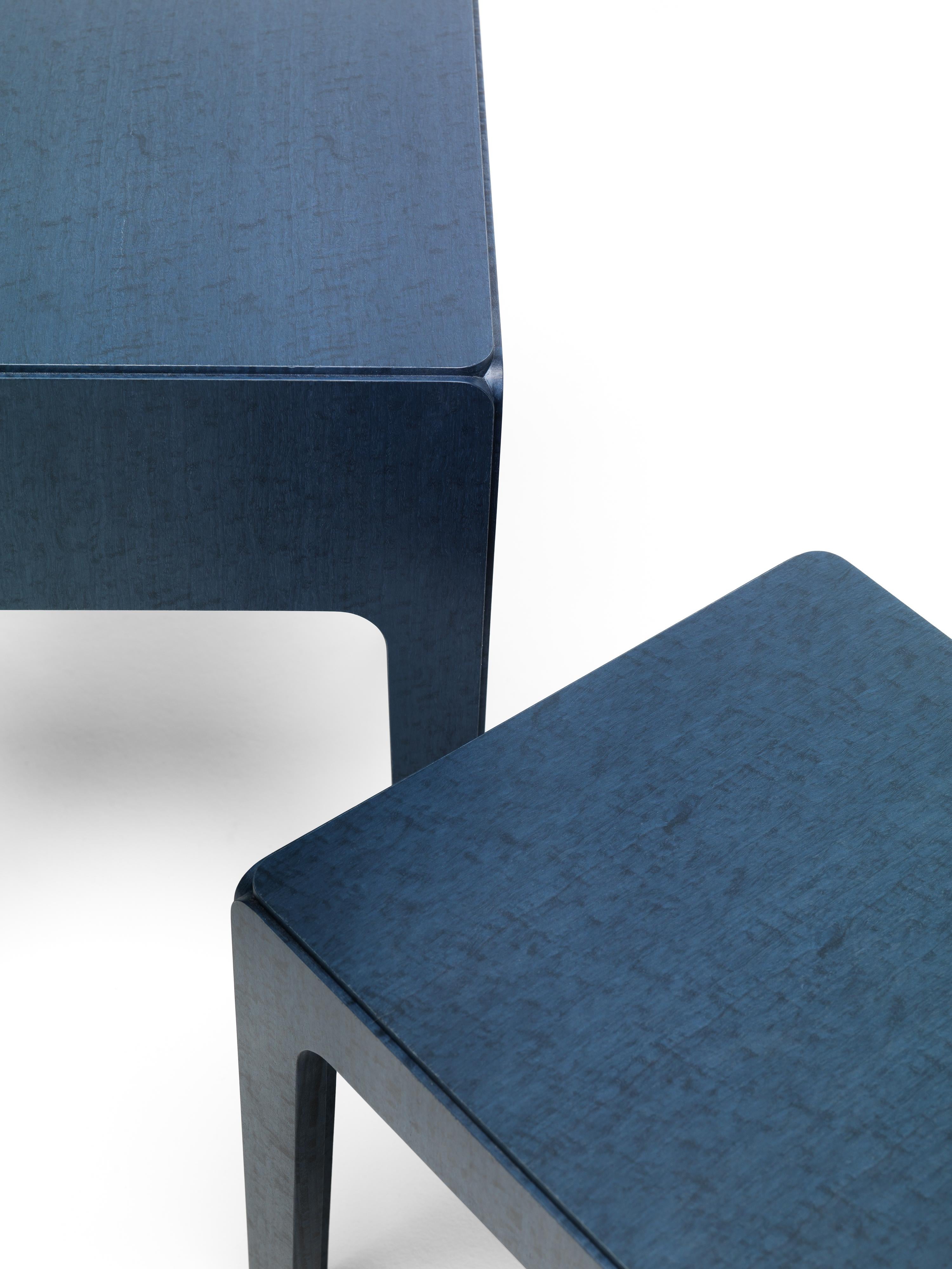 Eine Familie von Tischen mit 4 Beinen aus MDF, die mit blau gebeiztem Eukalyptusfurnier verkleidet sind. Durch einen einfachen Trick erscheint der Tisch, der in Wirklichkeit aus fünf Teilen besteht, wie aus einem massiven Block gemeißelt. Die vier