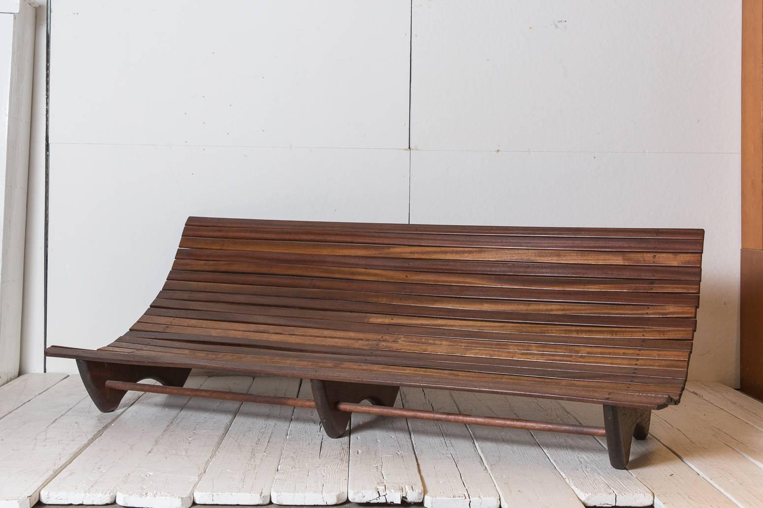 Wood stained Brazilian curved slatted bench with three legs.
