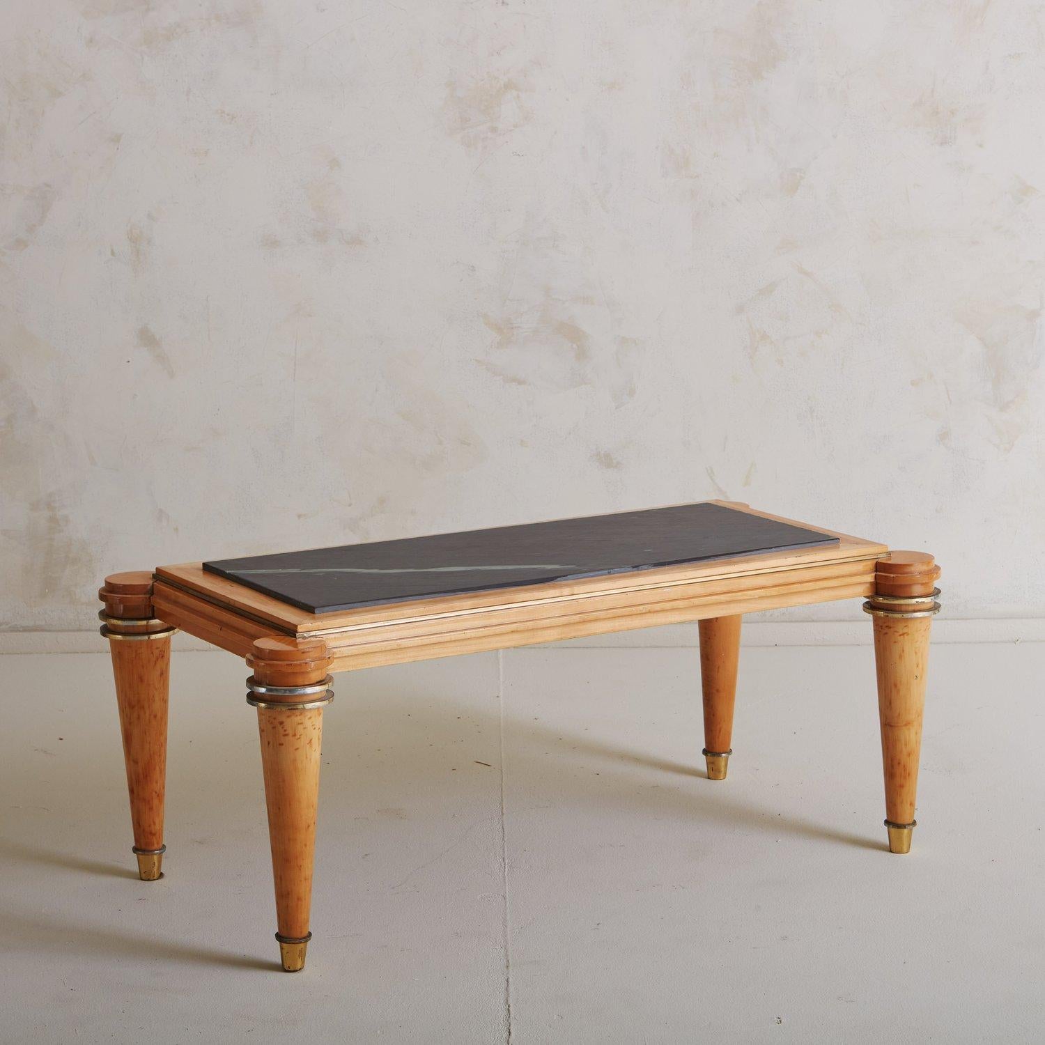 A 1940s Art Deco coffee table attributed to Etienne Kohlmann. This table has a rectangular wood frame and stands on four round, tapered legs with circular brass detailing and brass feet. It has a black stone tabletop that sits just above the wood