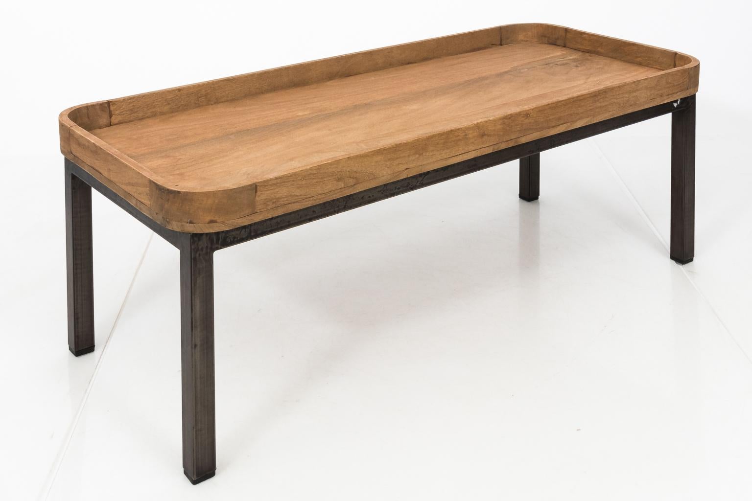 This coffee table is constructed from a large wooden tray with rounded corners resting on a handmade iron base. The wood has a natural matte finish.