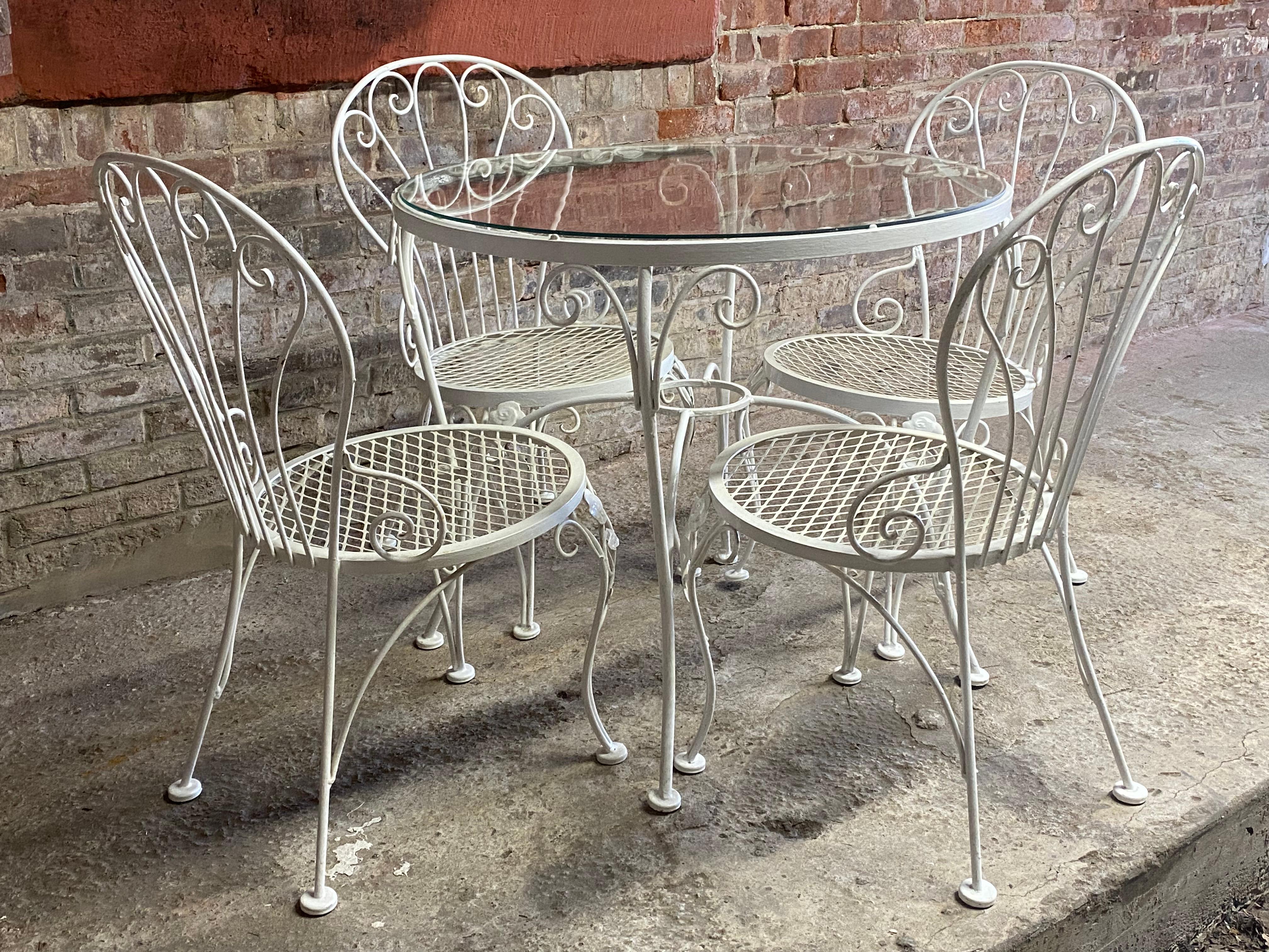 Woodard outdoor patio set that includes four scrolled chairs with mesh seats and a round glass top table. Circa 1965-70. Floral and scroll decoration. Good overall condition. Minor scratches to the glass top. Multiple coats of paint. Underside of