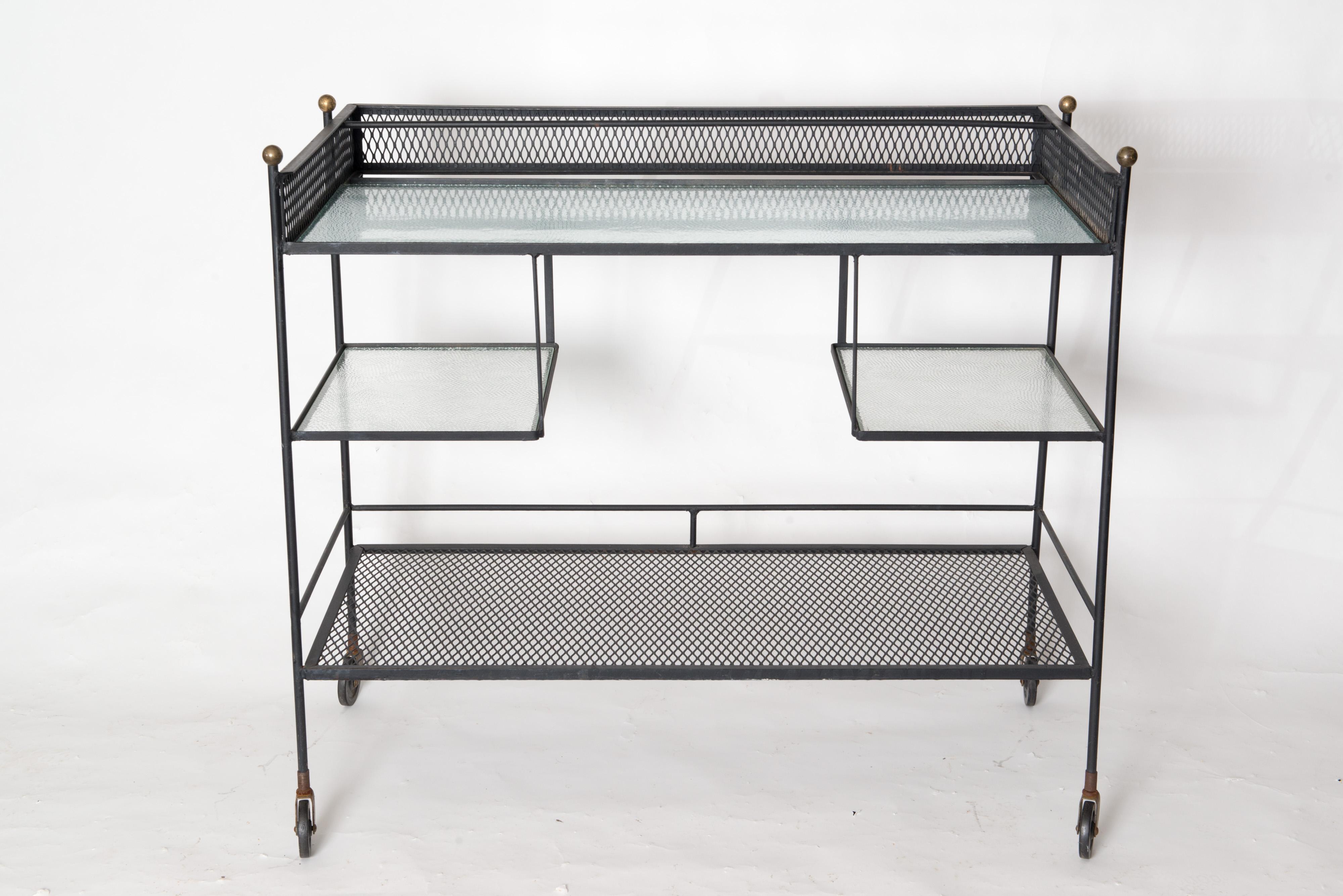This is a Mid-Century Modern three level wrought iron and glass rolling serving cart or bar cart with brass finials. The lowest shelf is metal mesh. The two middle shelves are glass. The top surface is glass. This cart is a very useful and fits in