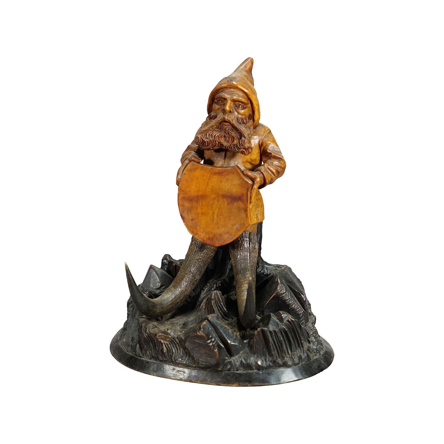 Wooden carved black forest dwarf sitting on a three stump.

A wooden handcarved dwarf holding a crest. Mounted on a wooden base featuring a three stump and rocks. The lindenwood design is manufactured in Switzerland, Brienz ca. 1900. 

The tradition