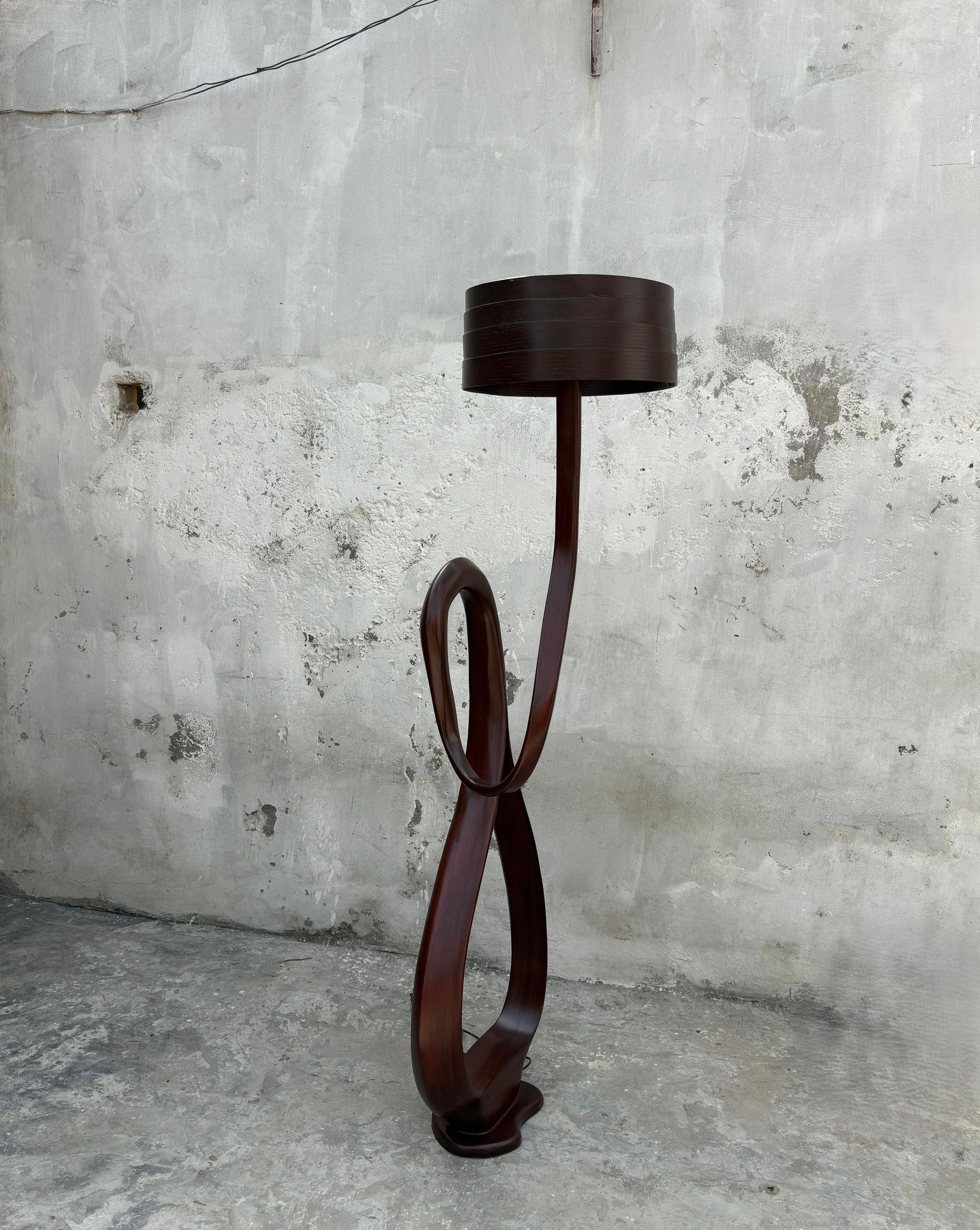 Die Stehlampe Nr. 6 aus der Fluentum-Serie ist eine skulpturale Leuchte, bei der die Bildhauertechniken dafür sorgen, dass sich der Körper der Leuchte bewegt: Das Holz ist oben schmaler und wird nach unten hin breiter, so dass eine Schleife