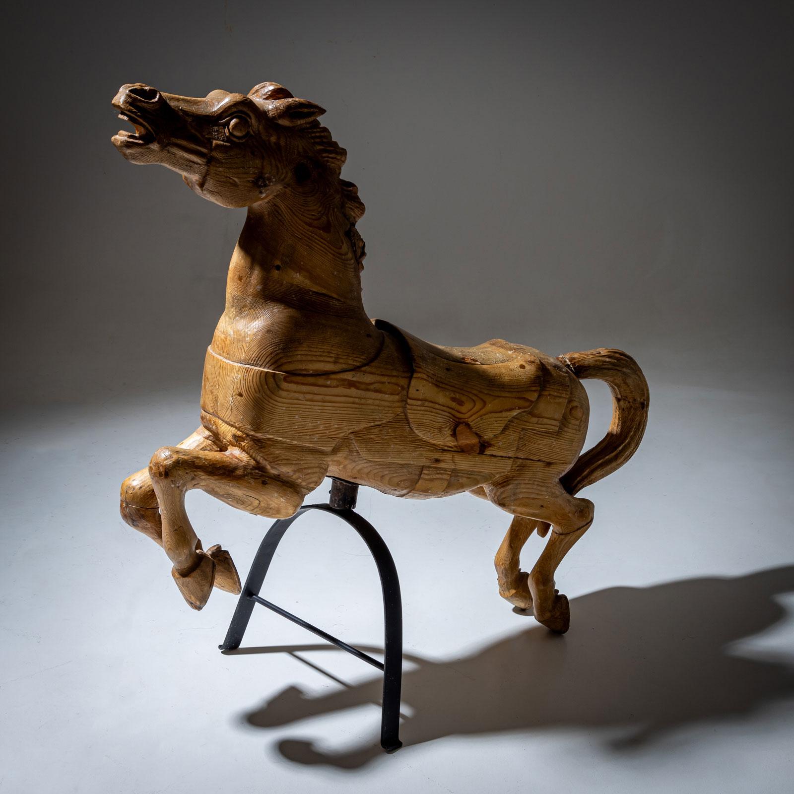 Grande cavallo da giostra in legno intagliato con cavalletto in ferro. La superficie in legno naturale intagliato con venature visibili gli conferisce un aspetto rustico. Il cavallo è in posizione rampante, il che gli conferisce dinamismo e charme.