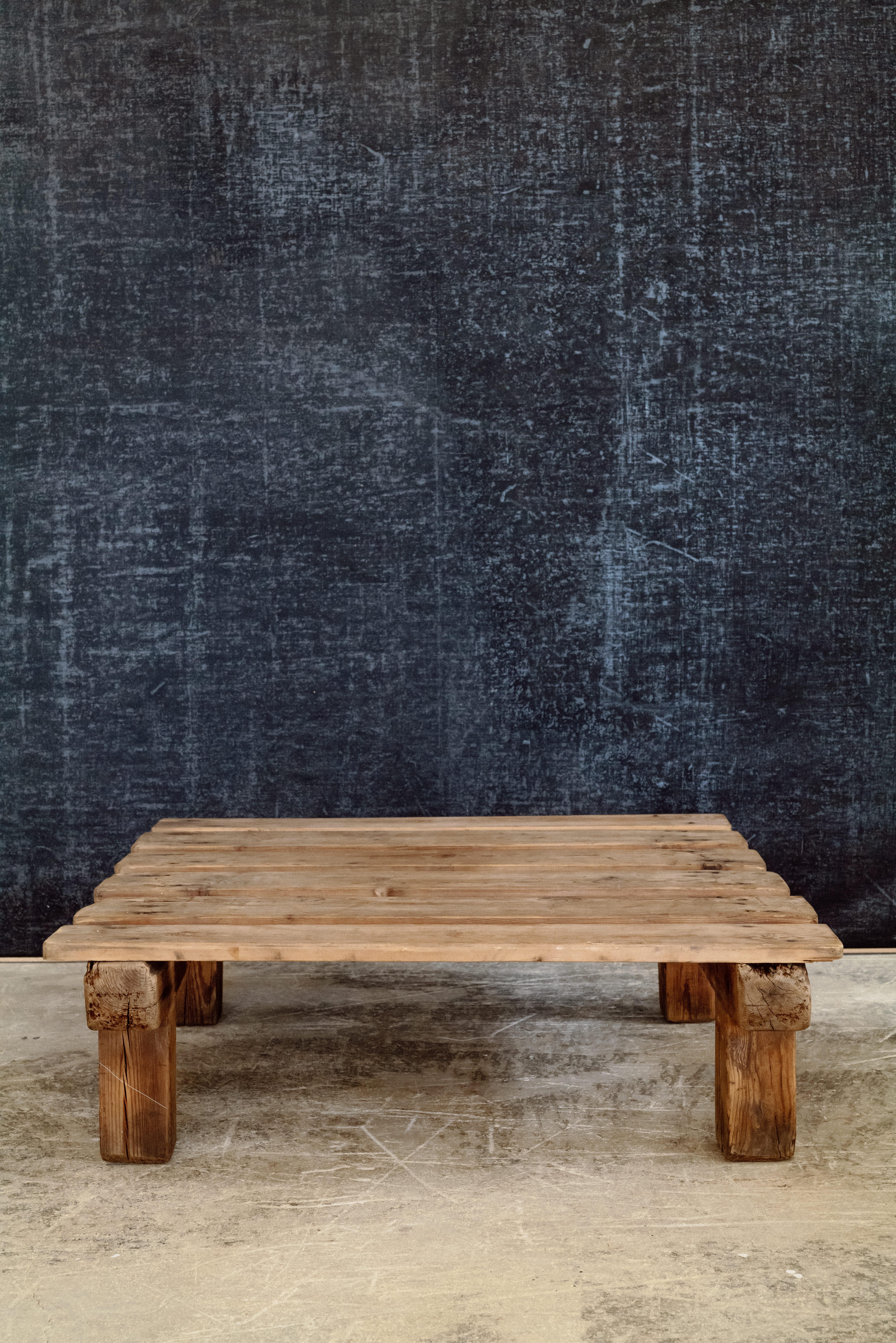 Une table basse fabriquée à la main à partir de lattes de bois récupérées, son design s'inspirant des palettes d'expédition utilitaires. La surface est composée de planches régulièrement espacées, créant un rythme texturé sur toute la surface. Les
