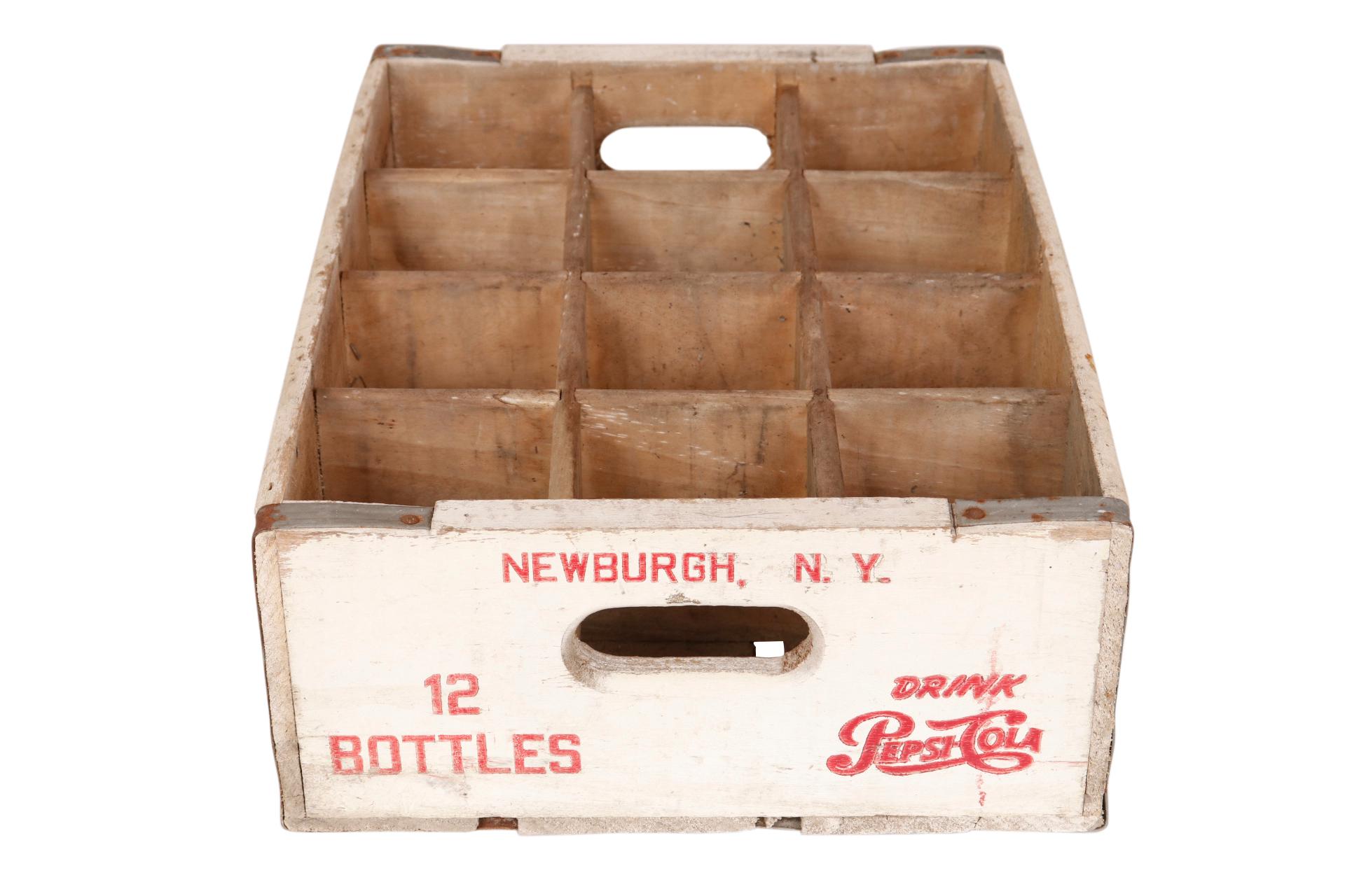 A 1950’s wooden Pepsi crate with a slat base. Reads “Drink Pepsi Cola” on each side in red lettering with “Newburgh NY, 12 Bottles” and oval cut out handles on the short sides. Metal straps support the corners and on the inside is stamped “Durable
