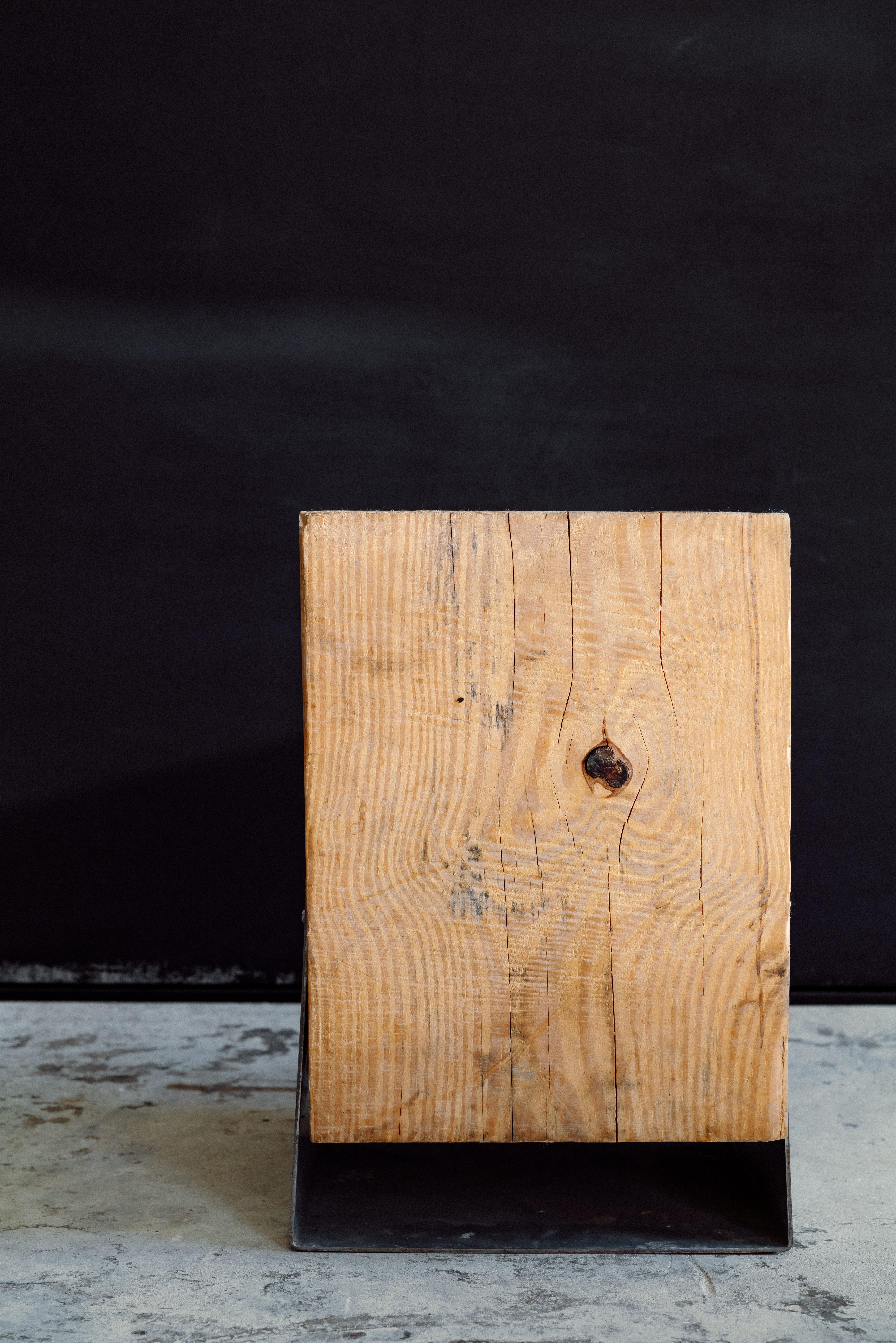 Un tabouret fonctionnel de style industriel fabriqué à partir d'un bloc de pin massif, ancré avec une plaque d'acier à la base pour plus de solidité et de stabilité. Le grain naturel du bois et l'usure visible contrastent avec la quincaillerie