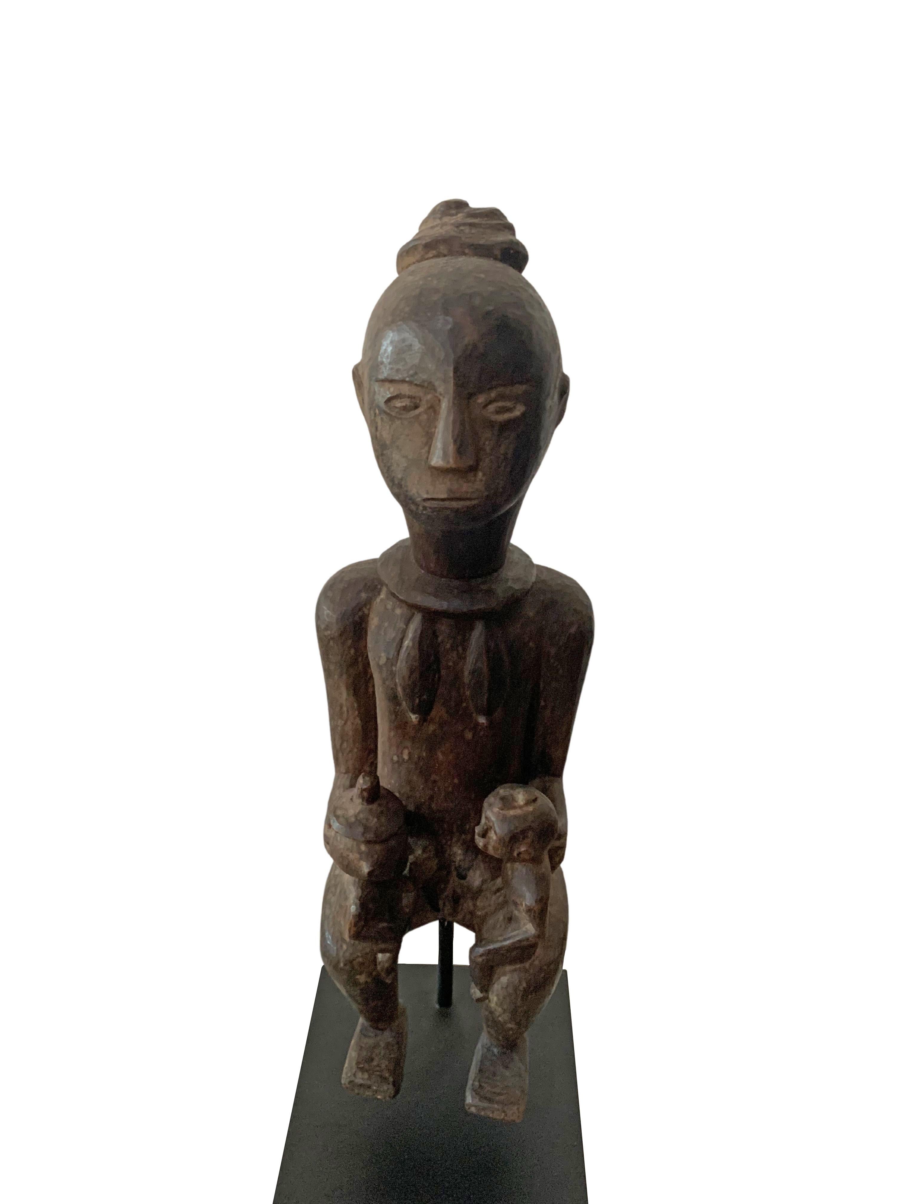 This wooden sculpture of a fertility figure originates from the Island of Sumba in Indonesia. It features subtle tribal engravings, and holds two child figures on each arm. Carved from a single block of wood the sculpture is mounted on a black iron