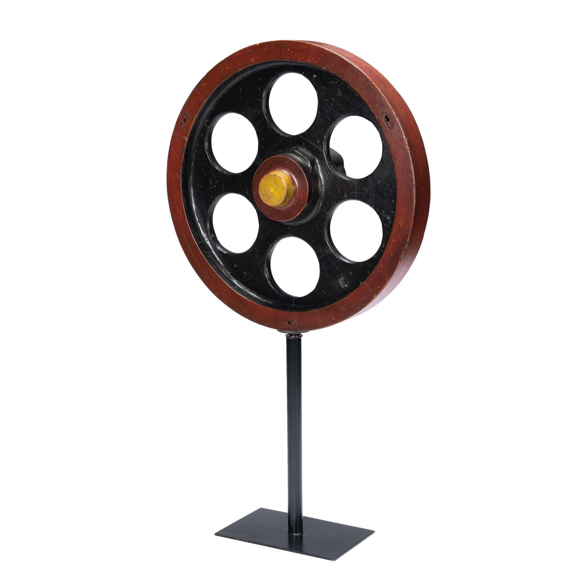 Wooden wheel casting mold with 6 circular holes around a raised axel stub. The wheel is painted in identifiable parts with areas of black, oxide red, and yellow. Mounted on a custom oxidized iron pedestal stand.