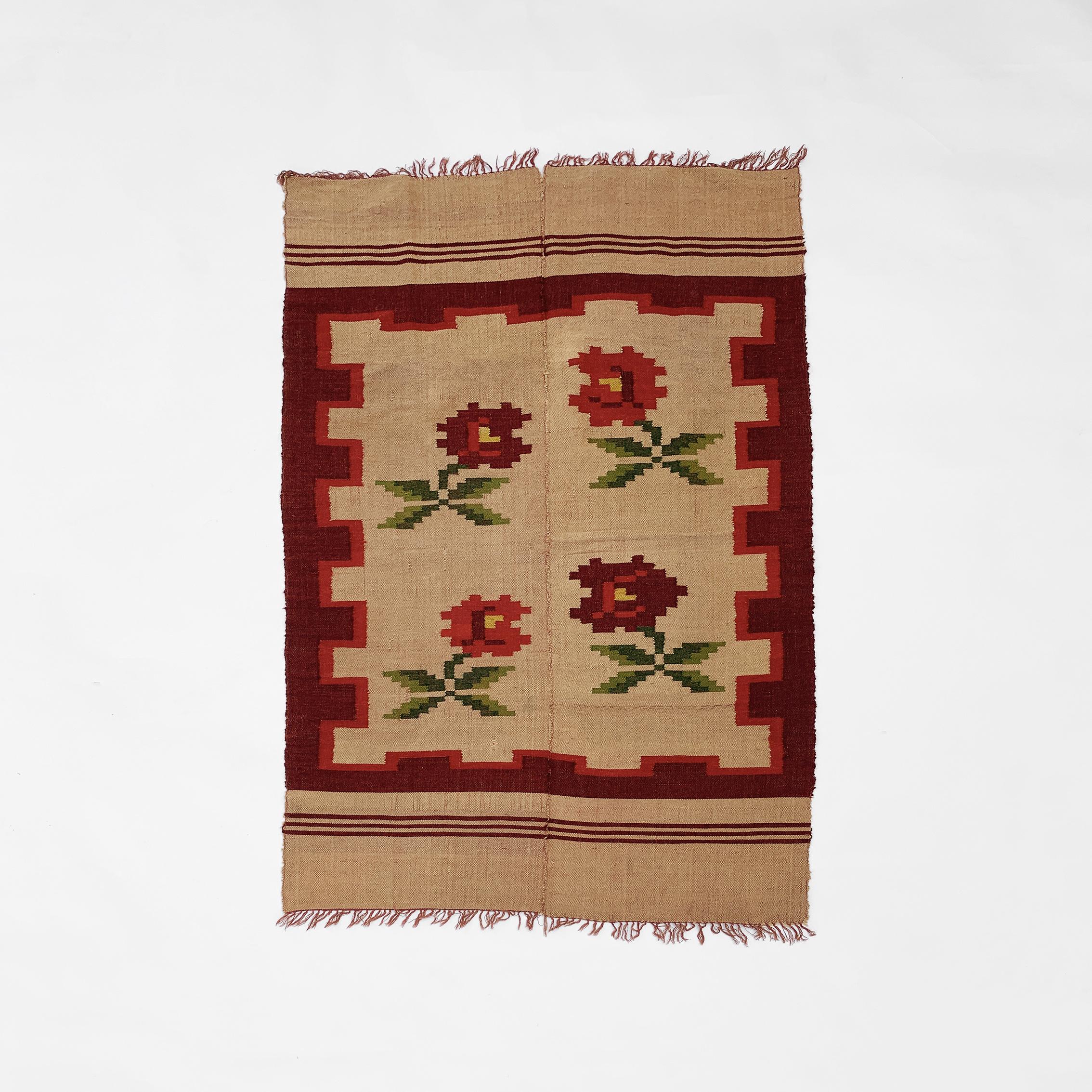 A small rug, imported from Greece, and featuring a red rose pattern on a tan background. This piece is handwoven from organic wool, and consists of two identically sized panels, hand-stitched together to create a larger rug. This technique was