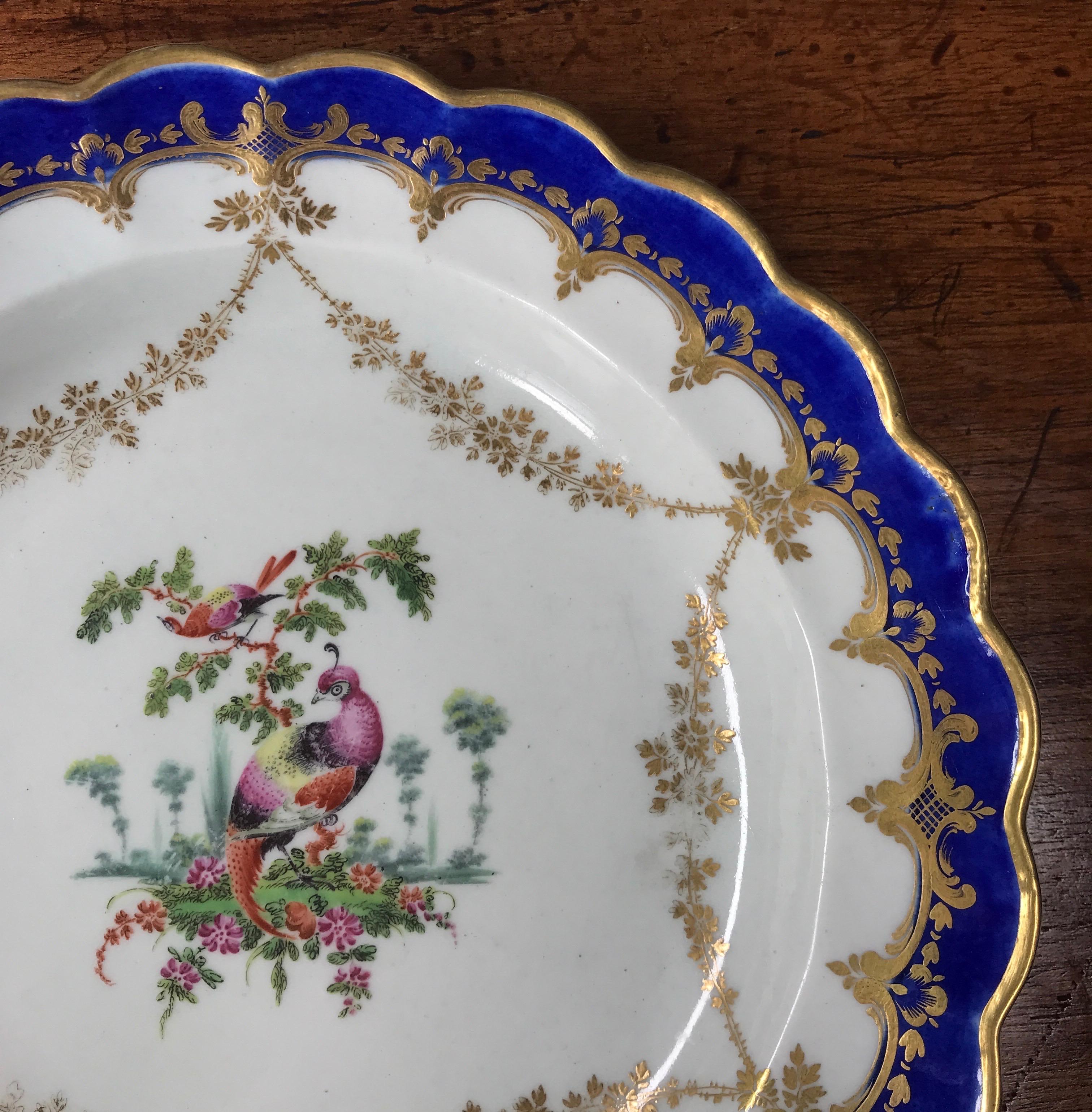 Worcester 'fancy birds' plate, painted to the centre with a small roundel of colourful birds, the rim with pendant swags of gilt foliage, a mazarin blue band to the scallops with further gilt scroll embellishments. 

Blue 'W' mark, circa 1770.