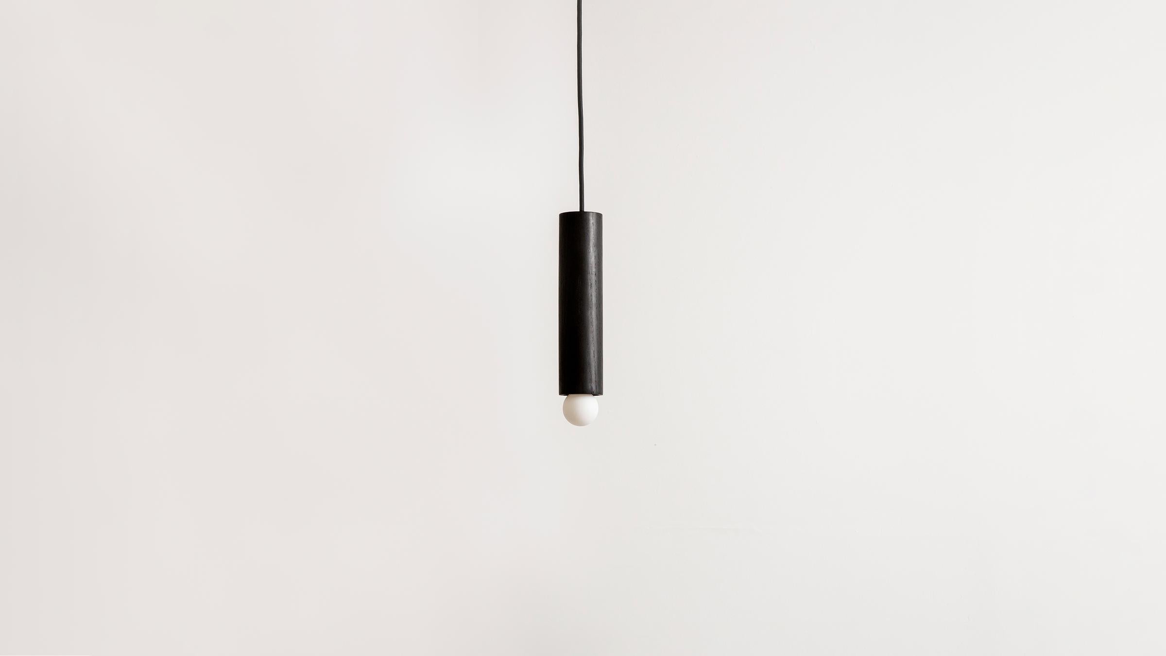 The lodge cord pendant small is crafted from a hand-turned oak dowel. Suspended from a cloth cord, the wooden dowel is a tactile armature punctuated by a single glowing bulb. Available in natural or oxidized oak with hewn brass or bronze details.