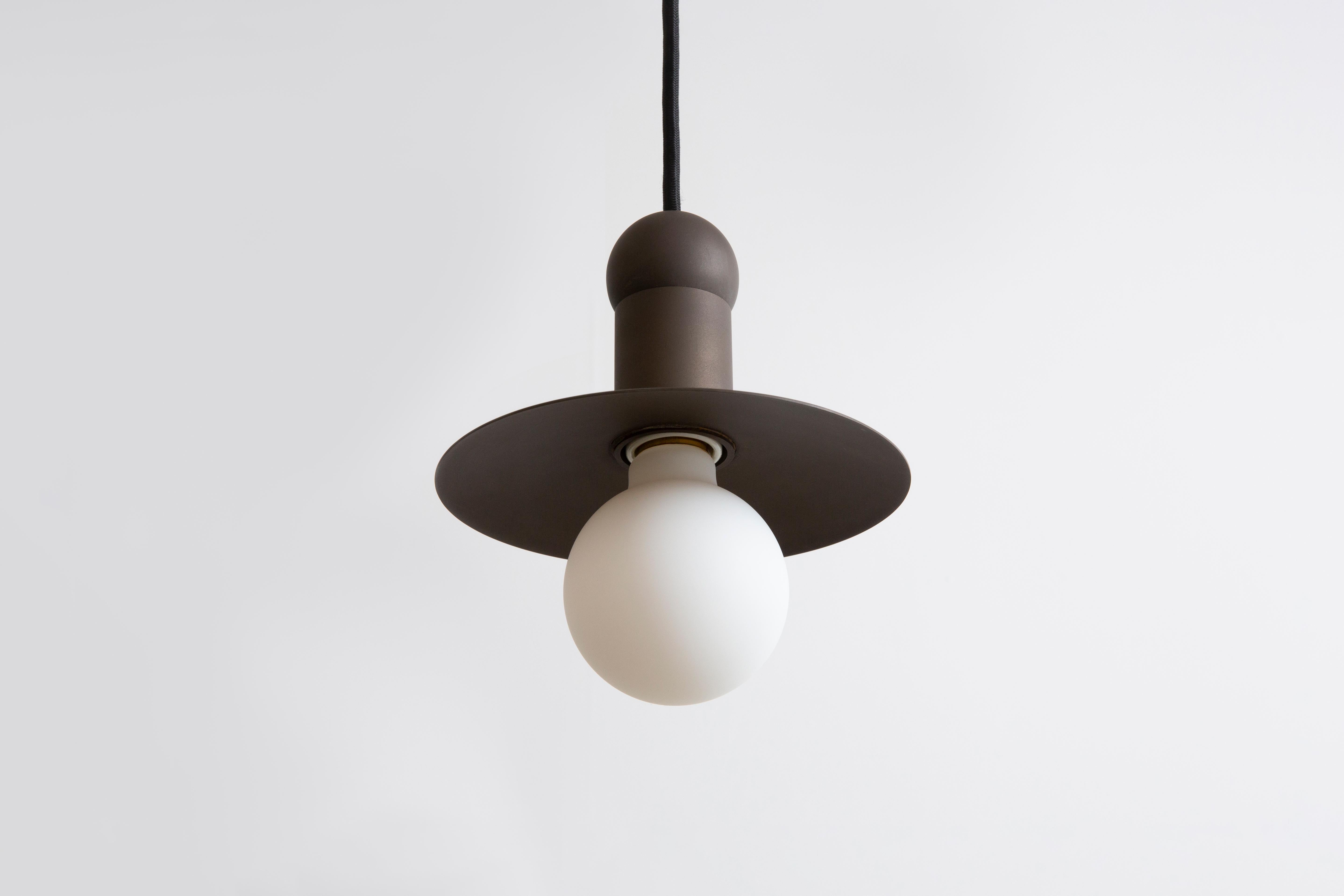 L'ORBIT CORD PENDANT place une seule ampoule incandescente contre une coupelle rayonnante stationnaire, explorant l'échelle et l'illumination d'une bougie des débuts de l'Amérique à travers la forme d'une suspension traditionnelle à éclairage vers