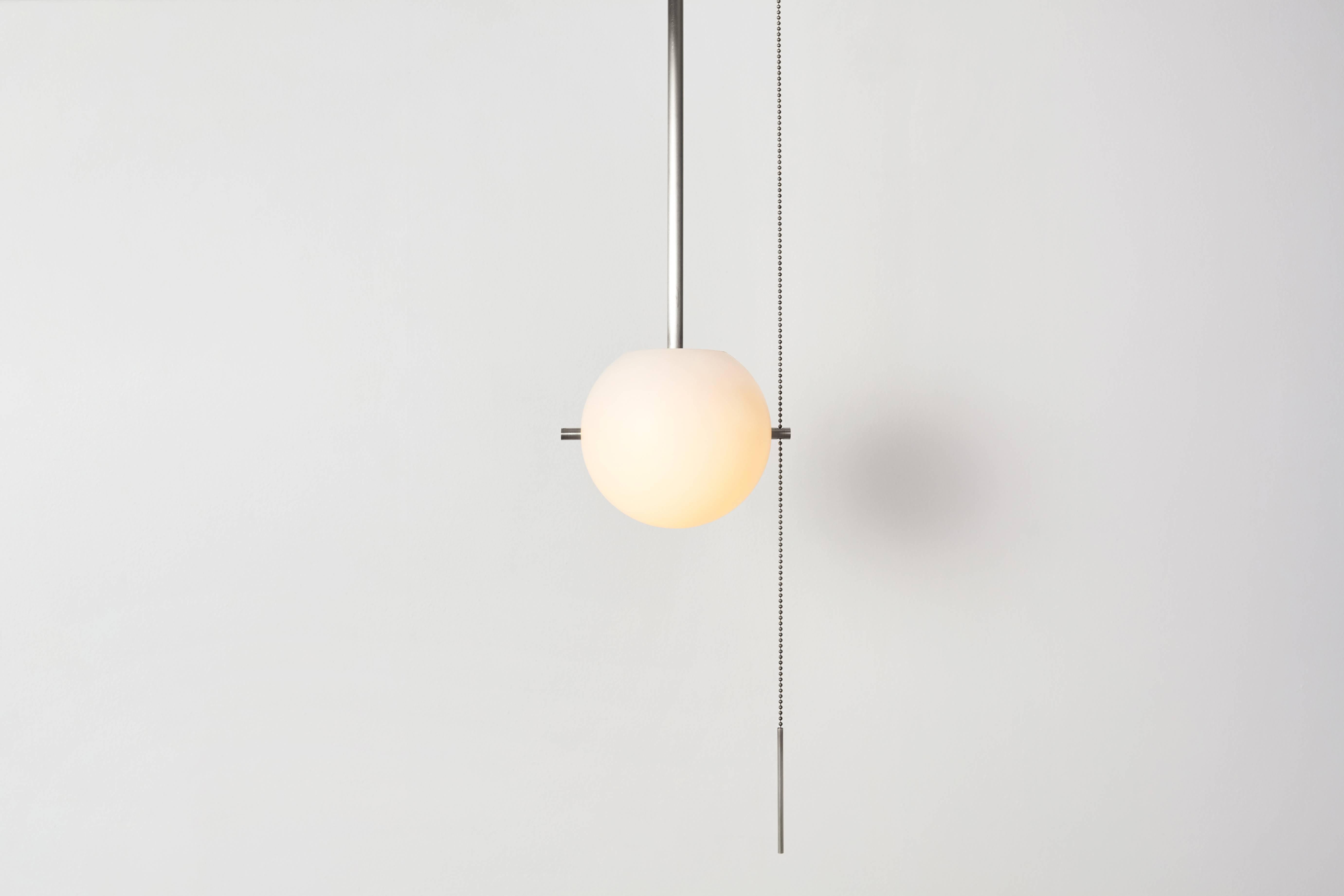 The SIGNAL FLUSH MOUNT features a hand-blown glass globe suspended from a luminous metal canopy by a pair of metal pins. A sinuous pull chain is weighted by a slender bar, giving the piece a jewel-like presence. Available as a flush mount or in