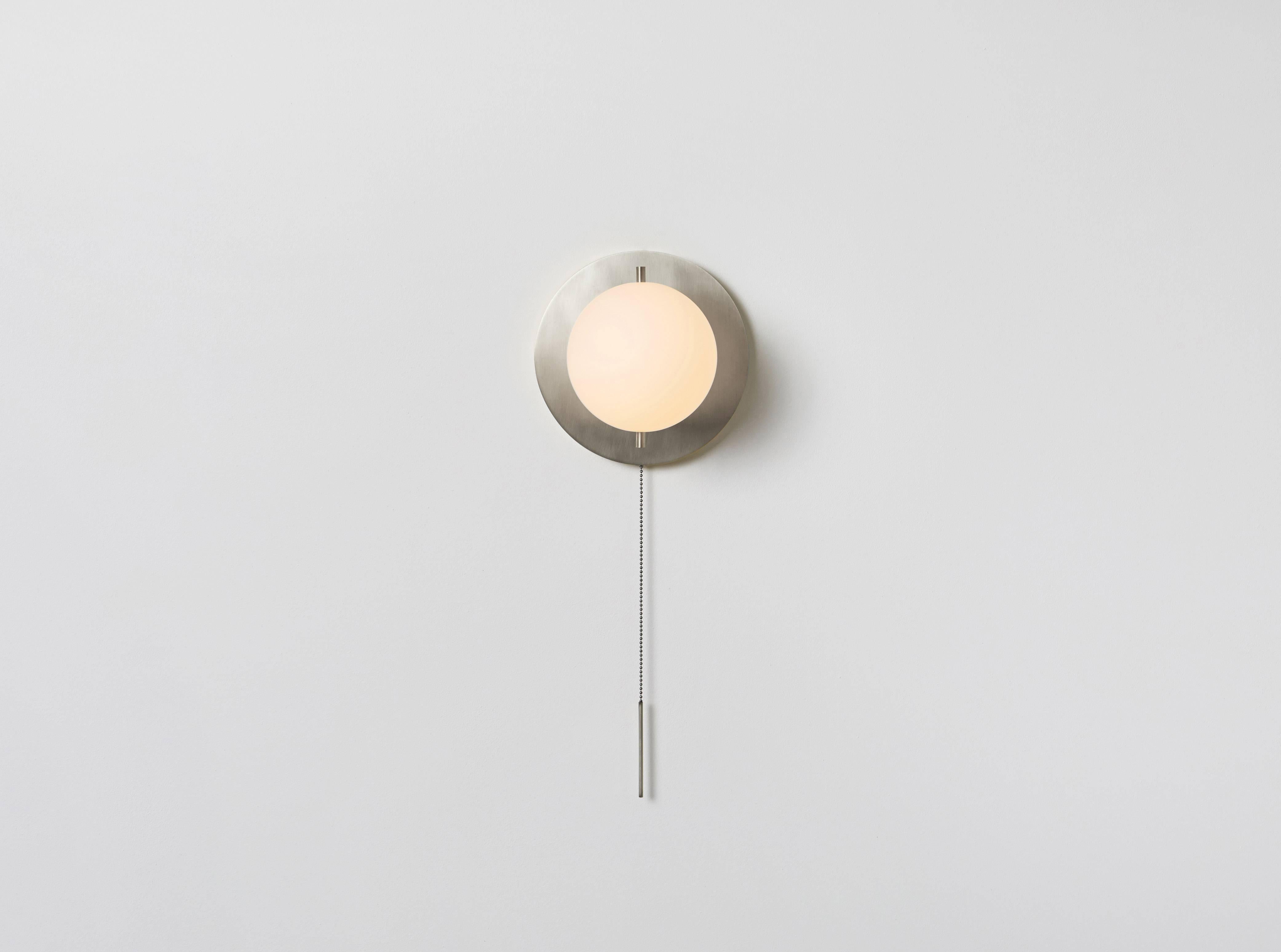 The Signal sconce features a glass globe. The fixture is delicately mounted between two metal pins against a luminous canopy, creating a hieroglyphic composition. A pull chain holds a slender rod, giving the piece a jewel-like presence. Made in the