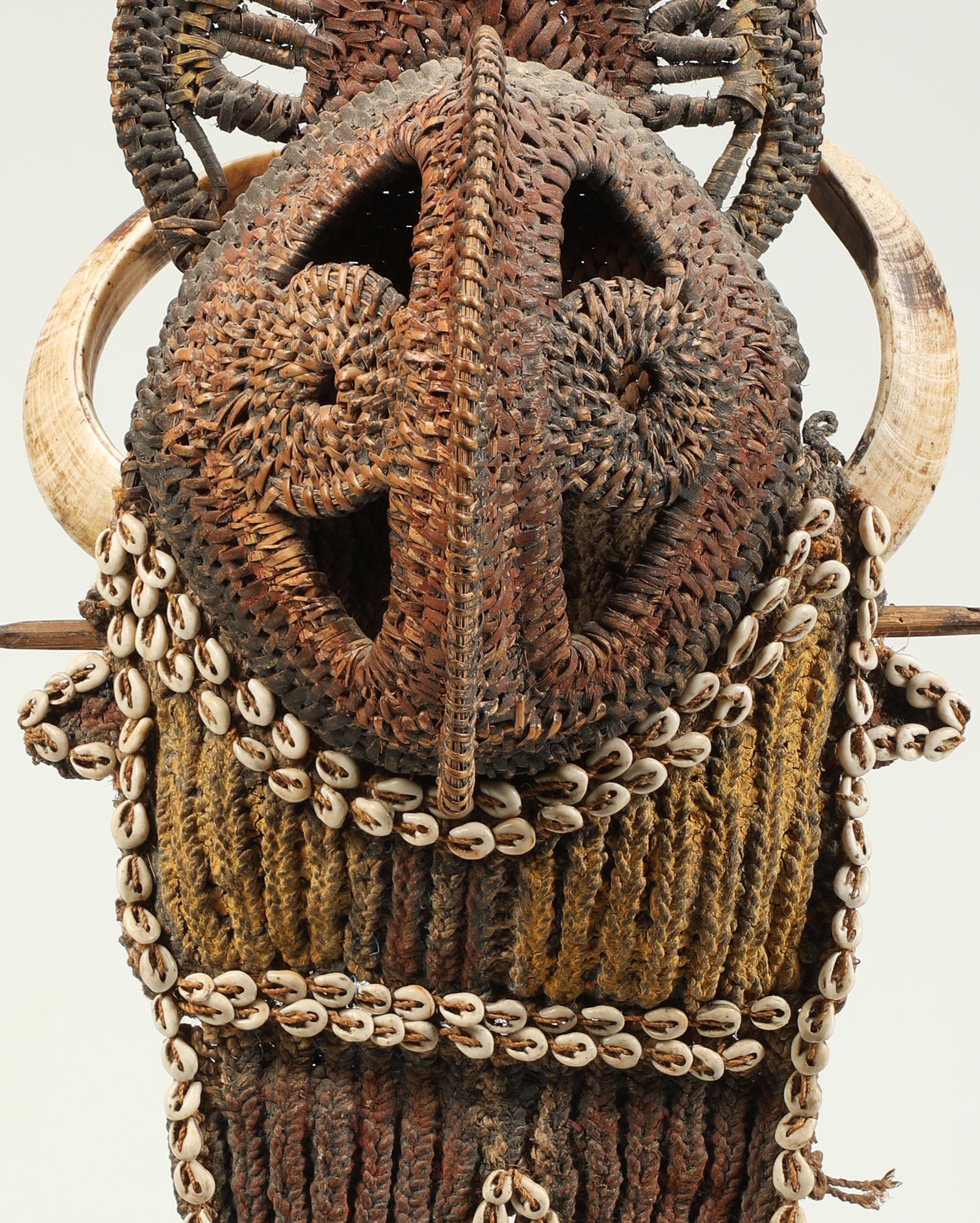 Woven basketry figural pectoral chest ornament with from Papua New Guinea. Woven yam mask style face with traces of red, black and yellow pigments, rings of attached cowrie shells and tusks.
On custom metal and wood base.
18 3/4 inches high, total
