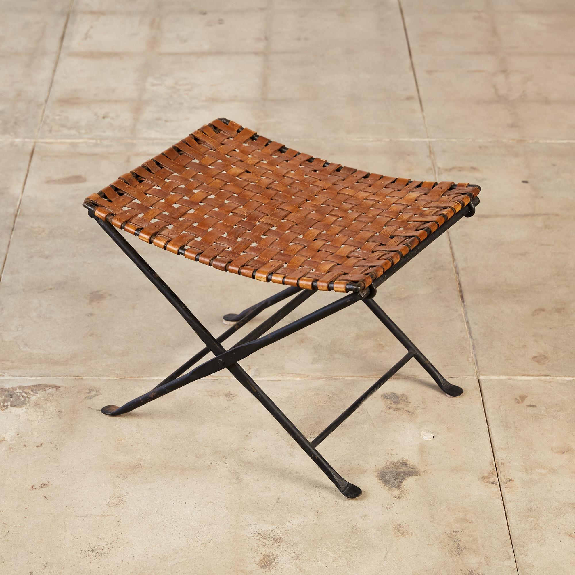 The stool features a folding wrought iron base with x-frame profiles with hammered metal details. The seat is composed of brown leather in a plain weave. The leather straps wrap around the metal frame showing off simplistic but fine
