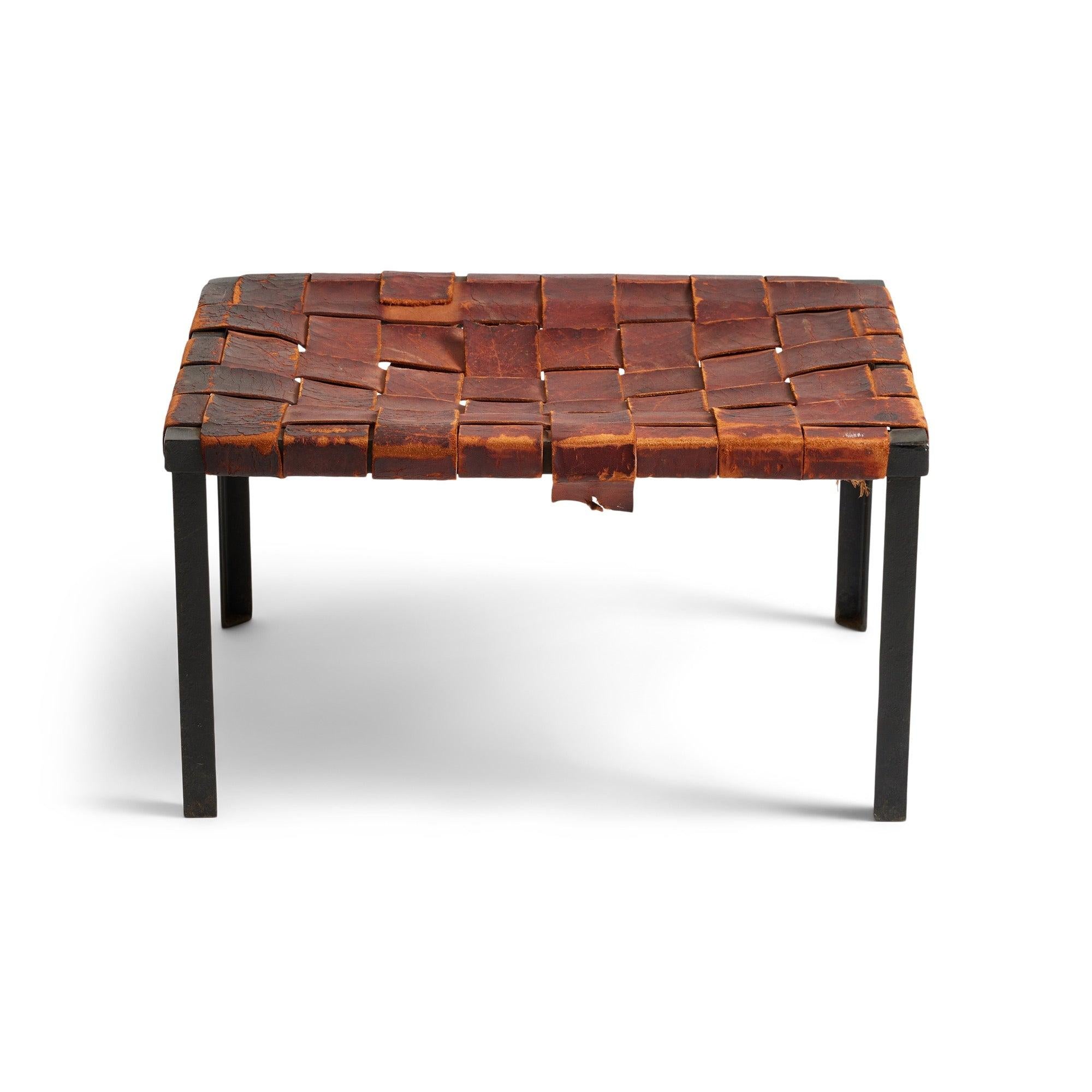 Un tabouret à quatre pieds en fer forgé, chaleureux et élégant, fabriqué à la main et conservant l'épaisse sangle de cuir cognac tissé d'origine.