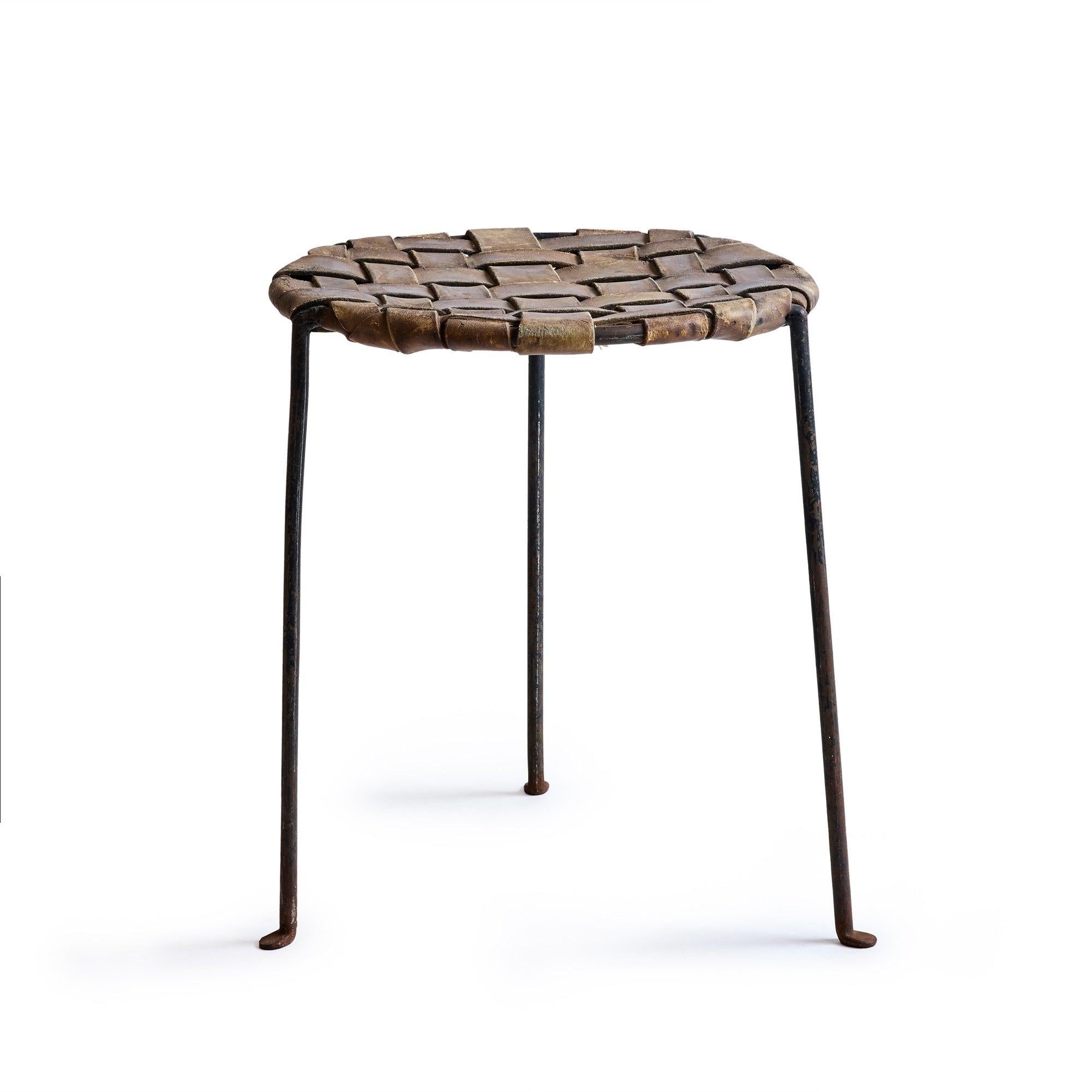 Un tabouret à trois pieds en fer forgé, chaleureux et élégant, fabriqué à la main et conservant l'épaisse sangle de cuir tissé cognac d'origine.