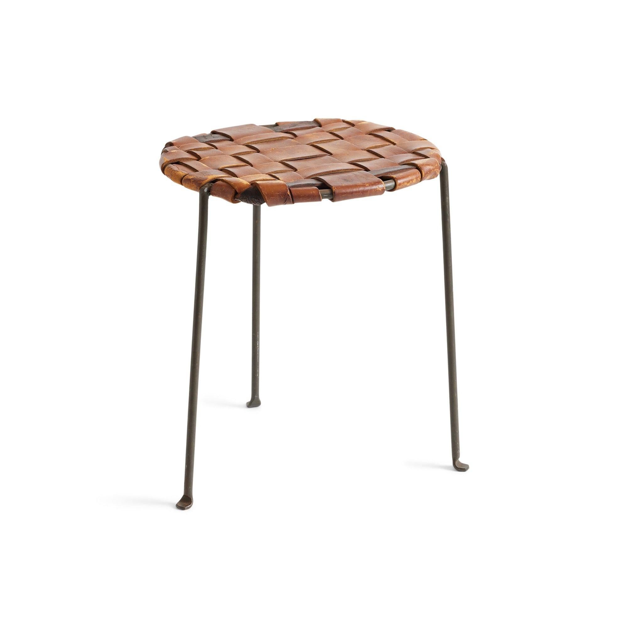 Un tabouret à trois pieds en fer forgé, chaleureux et élégant, fabriqué à la main et conservant l'épaisse sangle de cuir tissé caramel d'origine.