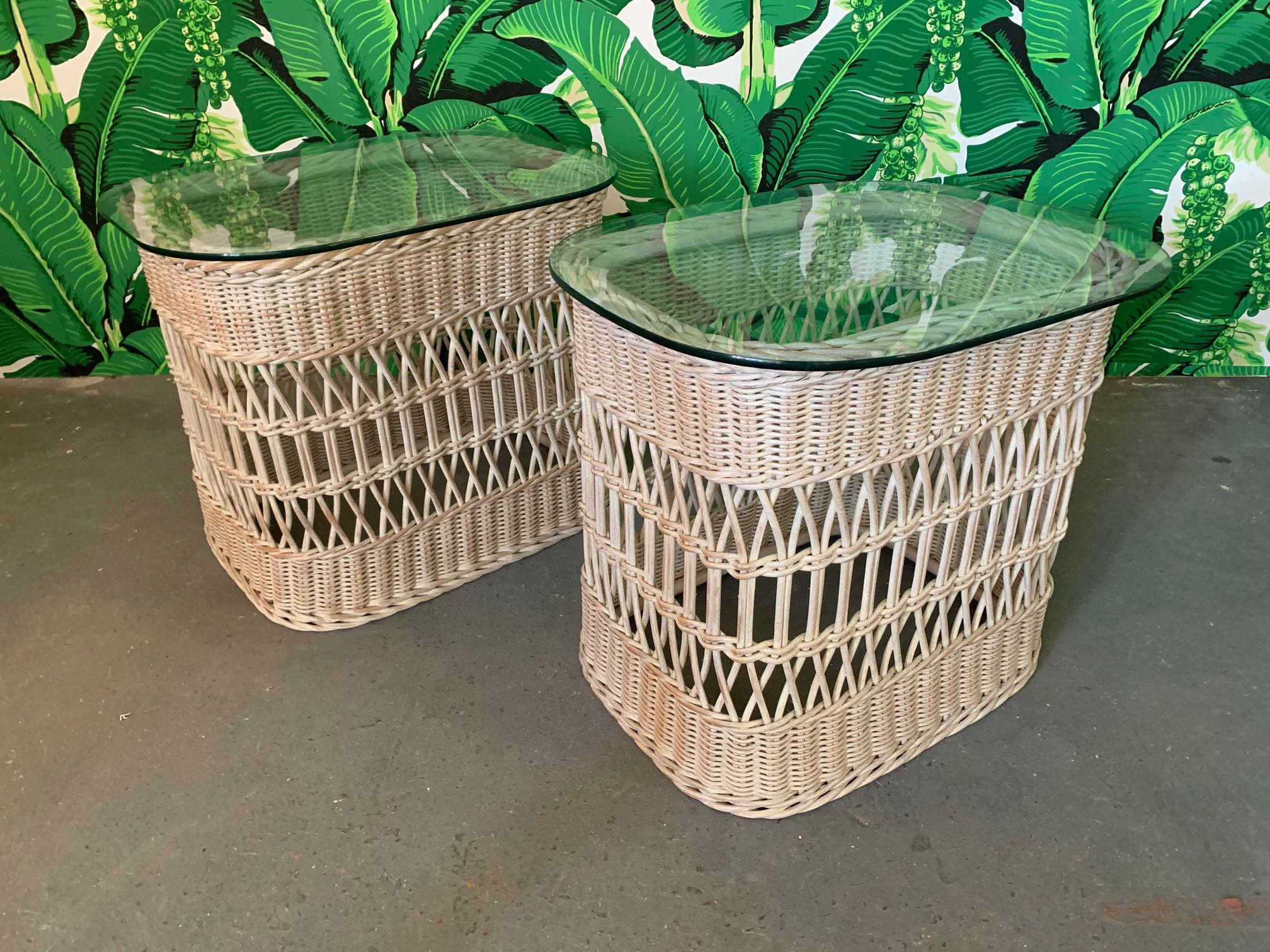 Pair of rattan side tables feature woven rattan and wicker base with glass top. Very good condition with only minor imperfections consistent with age. Bases are identical. One glass measures 26