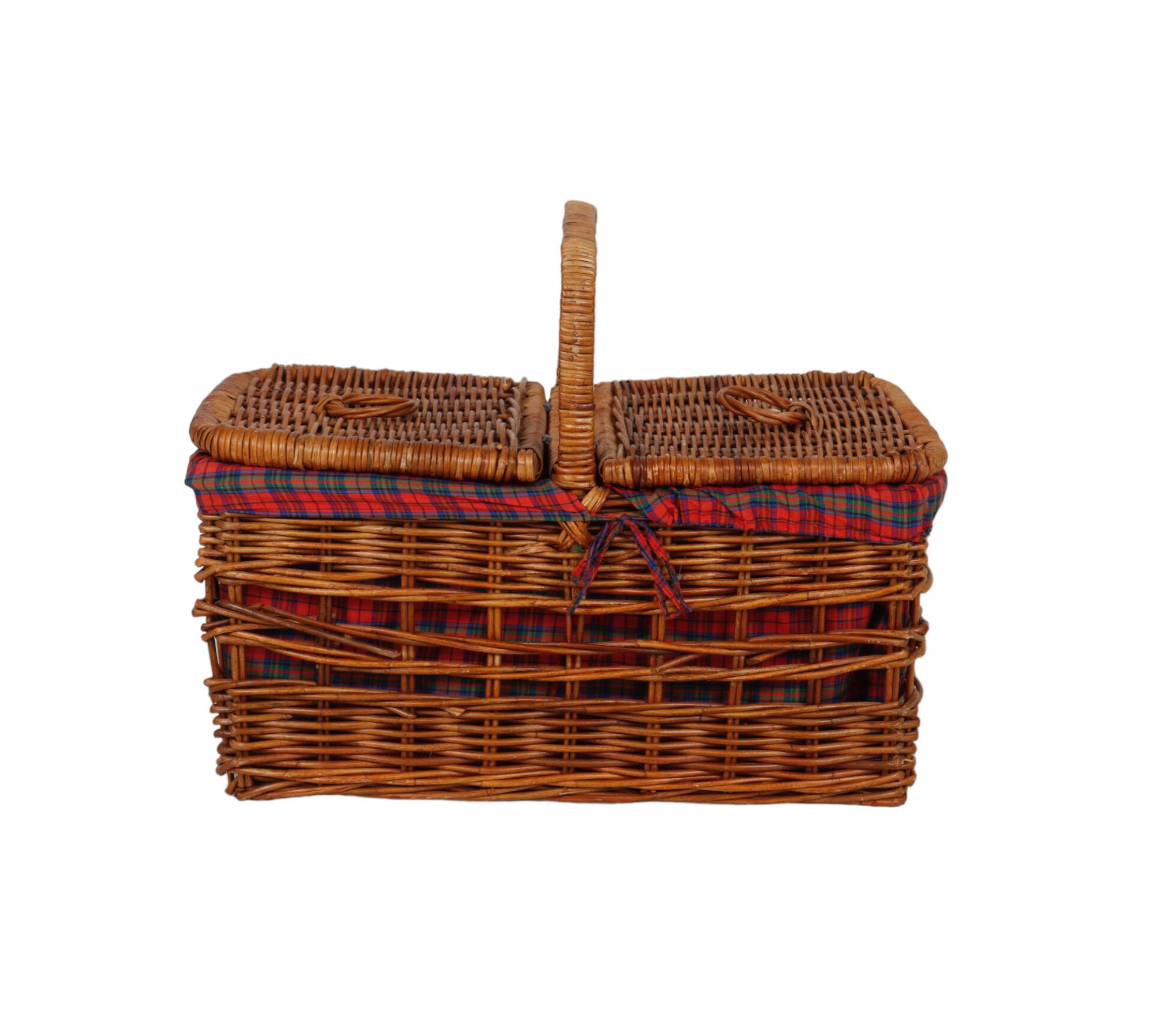 A large wicker woven rattan picnic hamper basket, with a plaid cloth lining and two handles. Two open weave bands decorate the sides allowing the pattern of the fabric inside to be seen. On top a single handle divides two lids, hinged at the center,