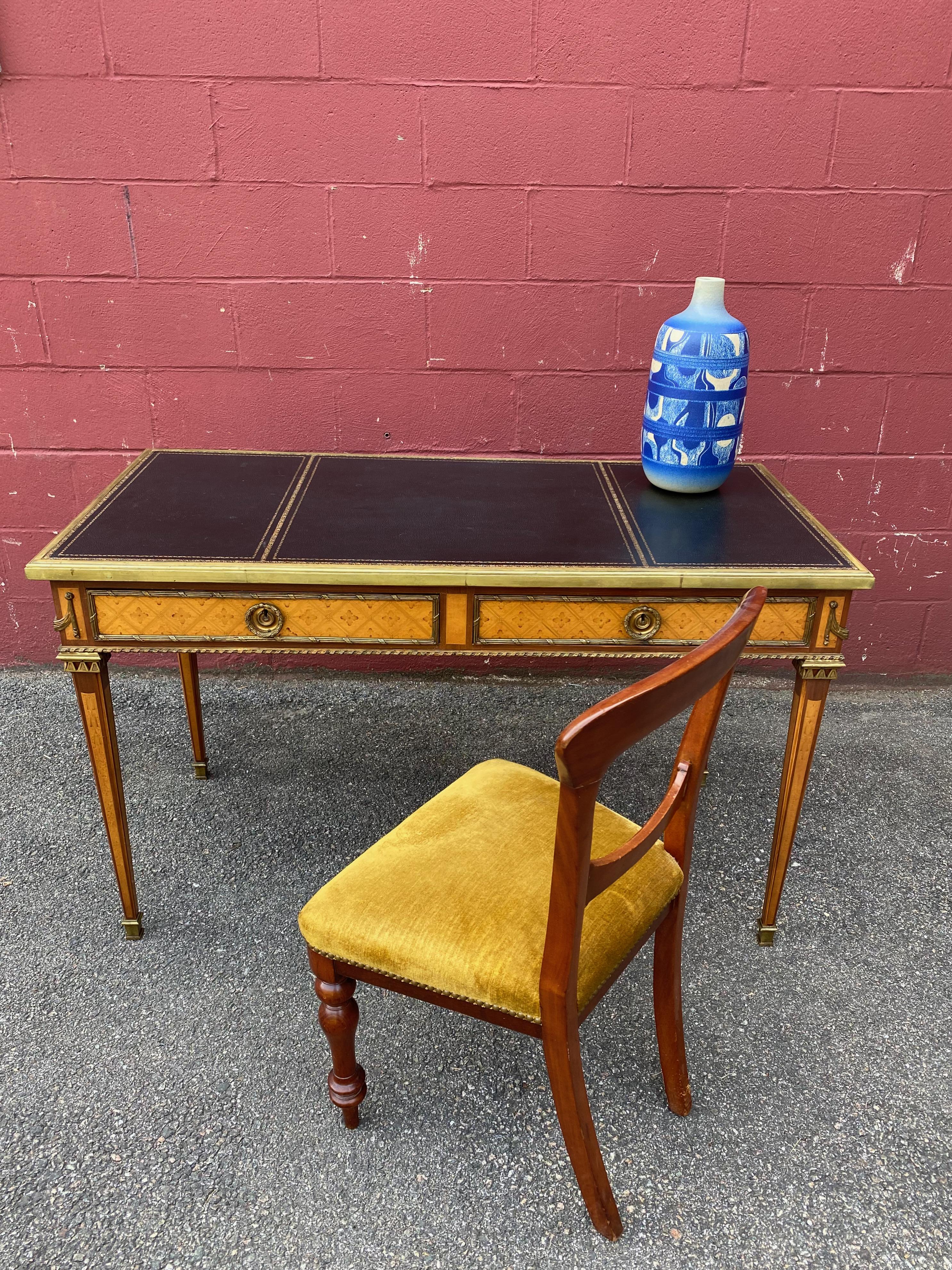 A small ladies writing desk with 2 drawers and pull out tablets on both ends. The black tooled leather top is surrounded by a stepped brass frame. The rich wood is a combination of satin wood and what appears to be mahogany with intricate inlays