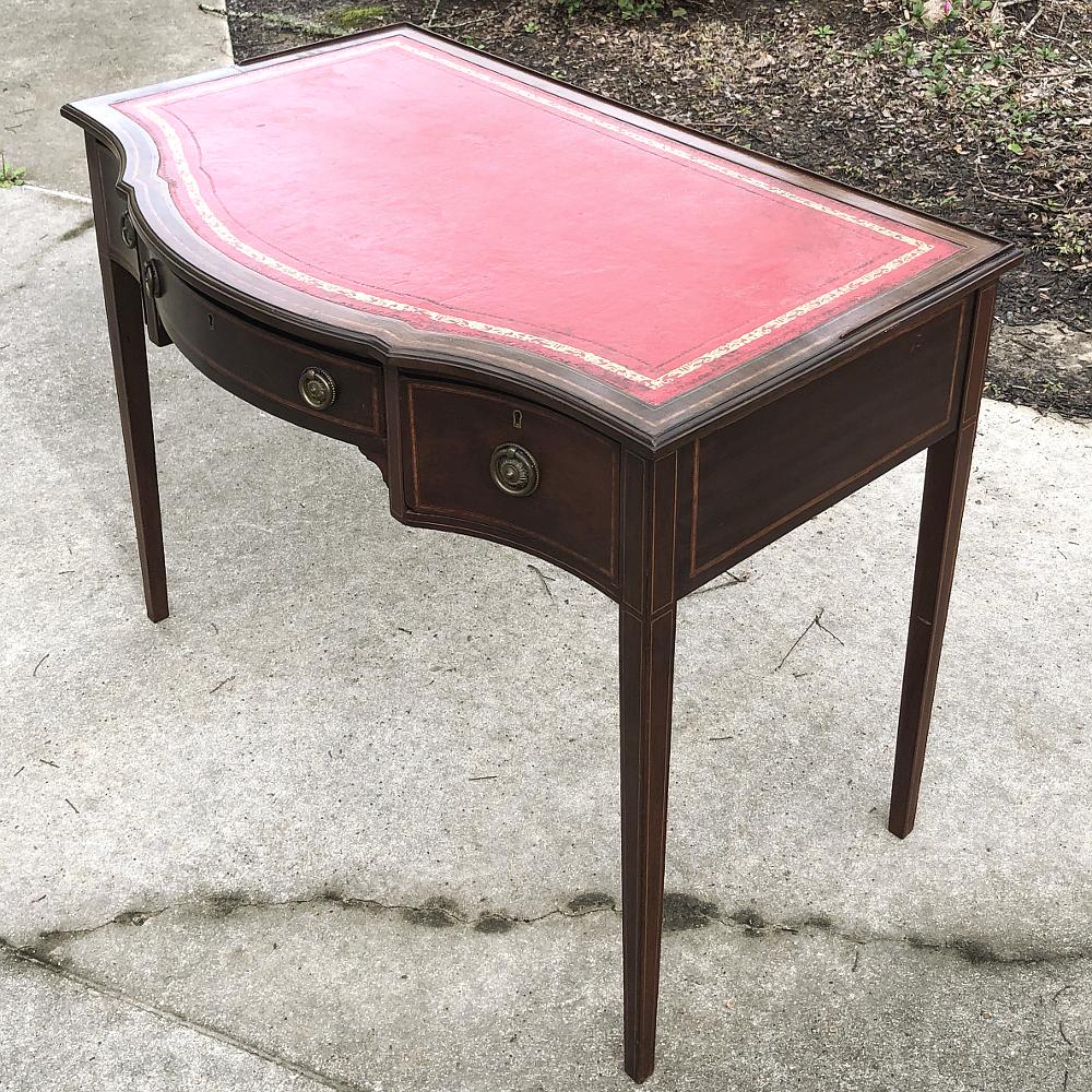 19th century English mahogany leather top writing table features its original tooled leather writing surface, brass pulls, and intriguing bowfront shape which is surprisingly ergonomic before the coining of the word itself! Handcrafted during the