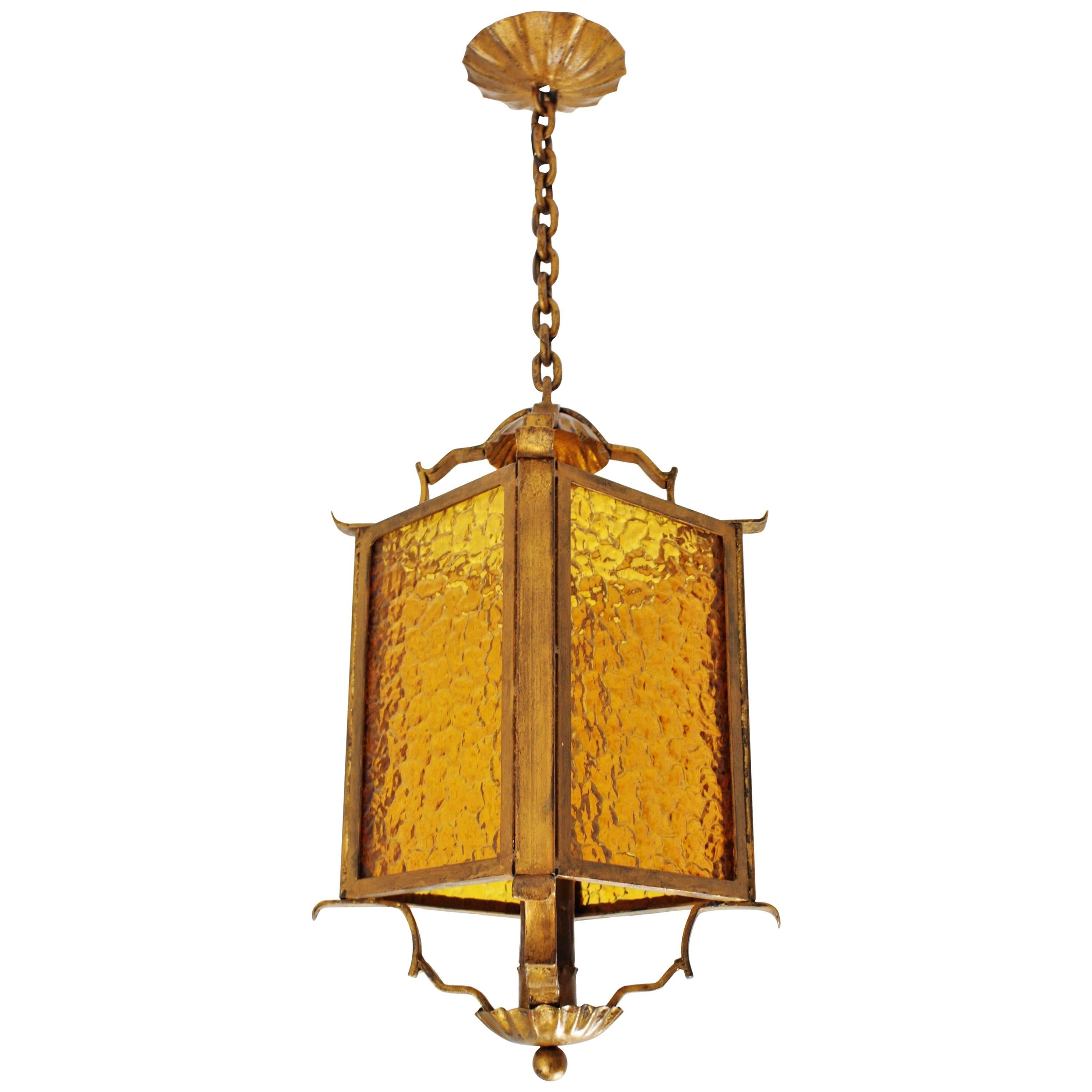 Sculptural gilt iron pagoda shaped pendant / lantern, Spain, 1930s.
This oriental hanging light pendant features a pagoda shape wrought iron chandelier. It has an structure with four sides comprised by a textured glass amber panel. It wears its