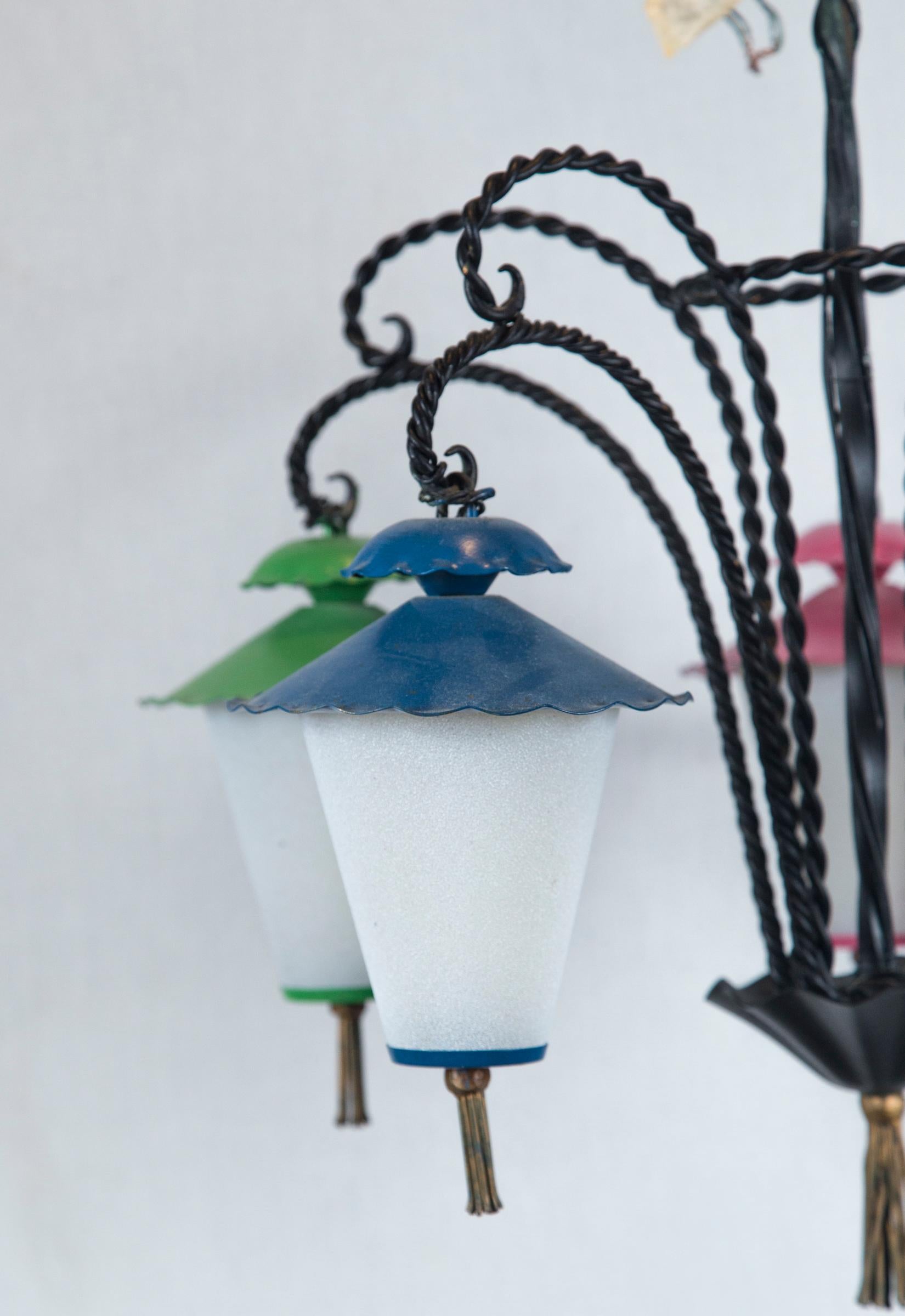 Araña de hierro forjado con cinco faroles colgantes de estilo japonés. Los faroles son de vidrio con leche, con la parte superior y la inferior de metal, adornadas con borlas de latón. Cada farol está pintado de un color diferente.