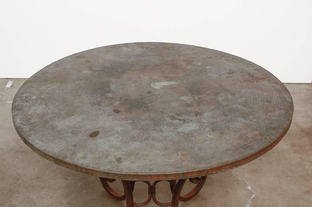 Dramatic scrolled wrought iron dining table featuring a hammered copper top. The copper top showcases a hand-hammered design in a concentric sunburst pattern. The wrought iron base is constructed from thick square iron bars with scrolled ends and