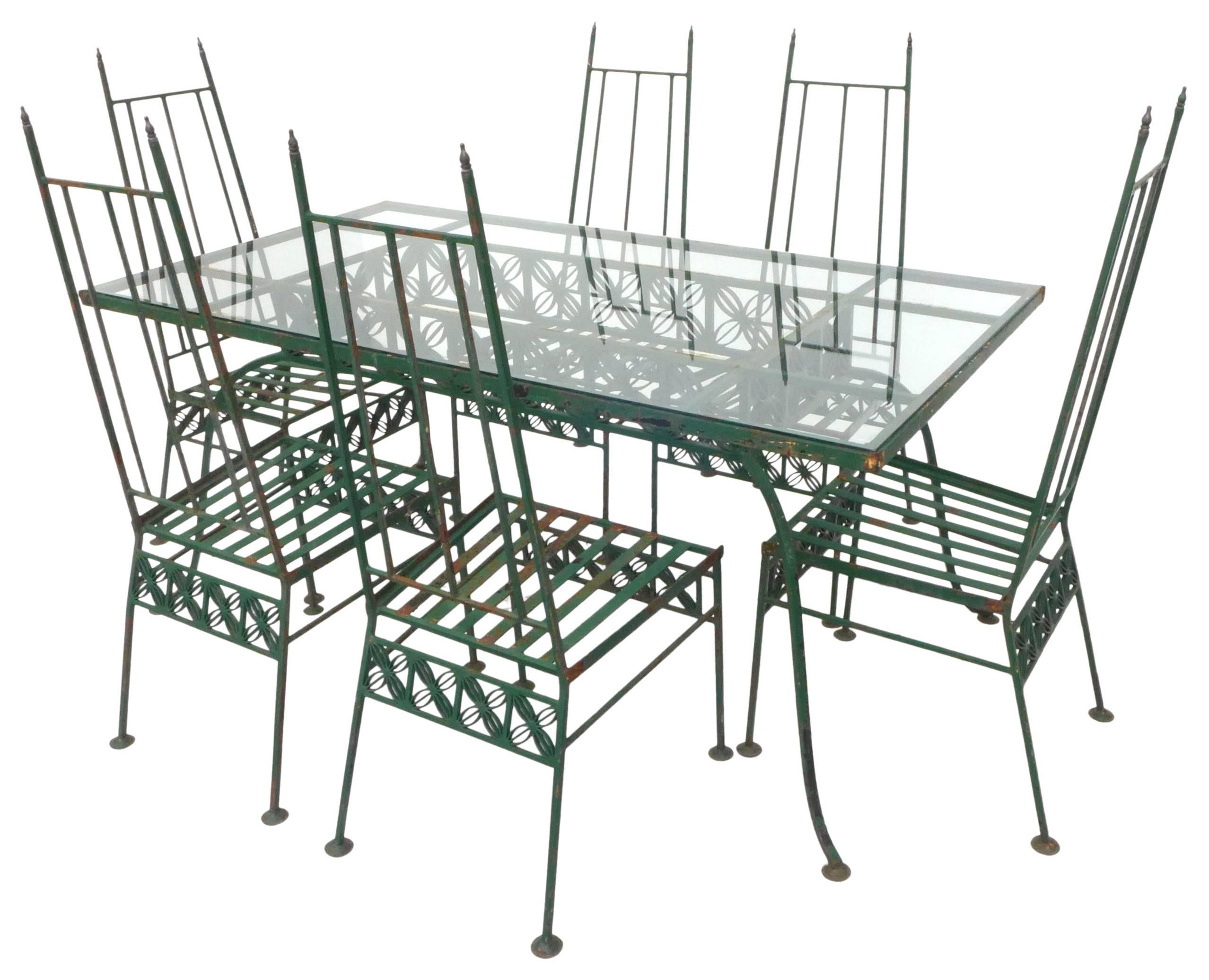 An incredible outdoor dining set for six in wrought iron and glass. A striking group with repeating, graphic, floral details stretched across each piece. High-back chairs sporting brass finials and disc feet. Painted in green with occasional