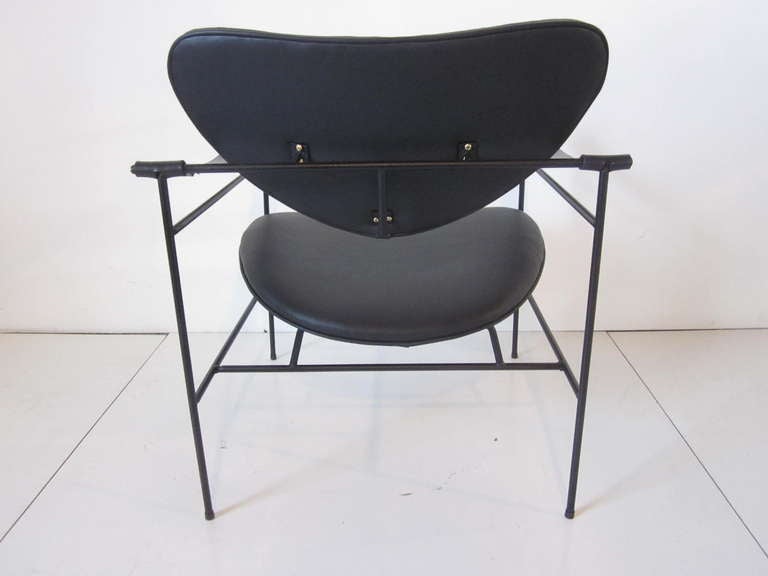 A black wrought iron framed lounge chair having black leather strap arms and brass hardware with the seat back and bottom upholstered in a soft and supple black leather. The back rest is arrow shaped giving the chair interest and great back support