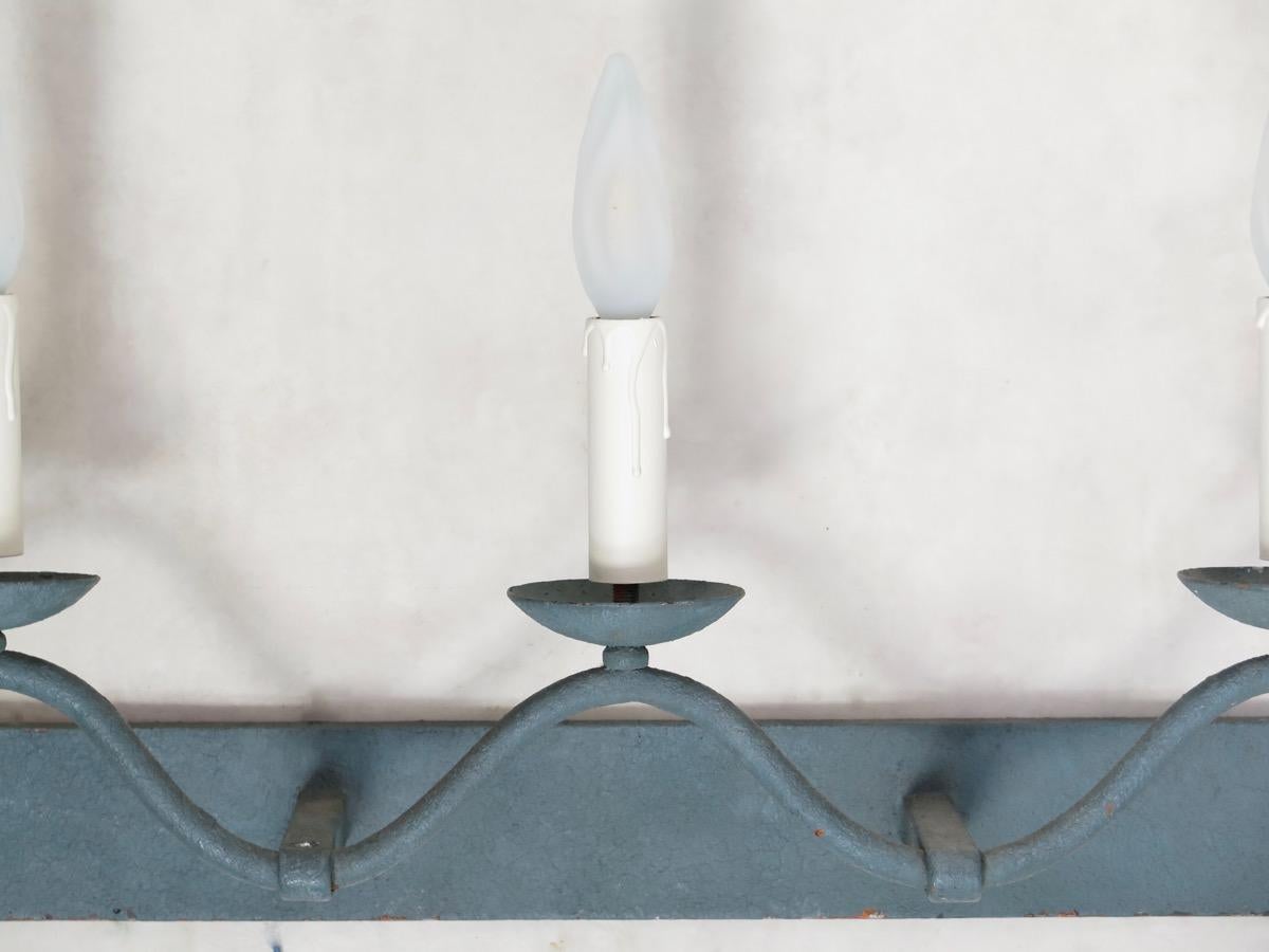 Applique à cinq lumières en fer forgé, longue et ondulée, peinte en gris/bleu, avec un apprêt orange visible en dessous.