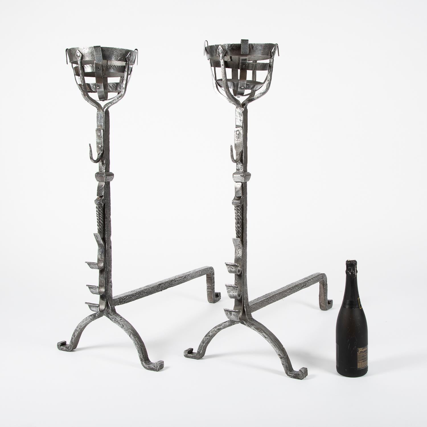 A pair of wrought iron cresset fire dogs, English, circa 1800.

Cresset tops used for the warming of flasks of wine, and the fronts each have three rests for fire tools or roasting spits.