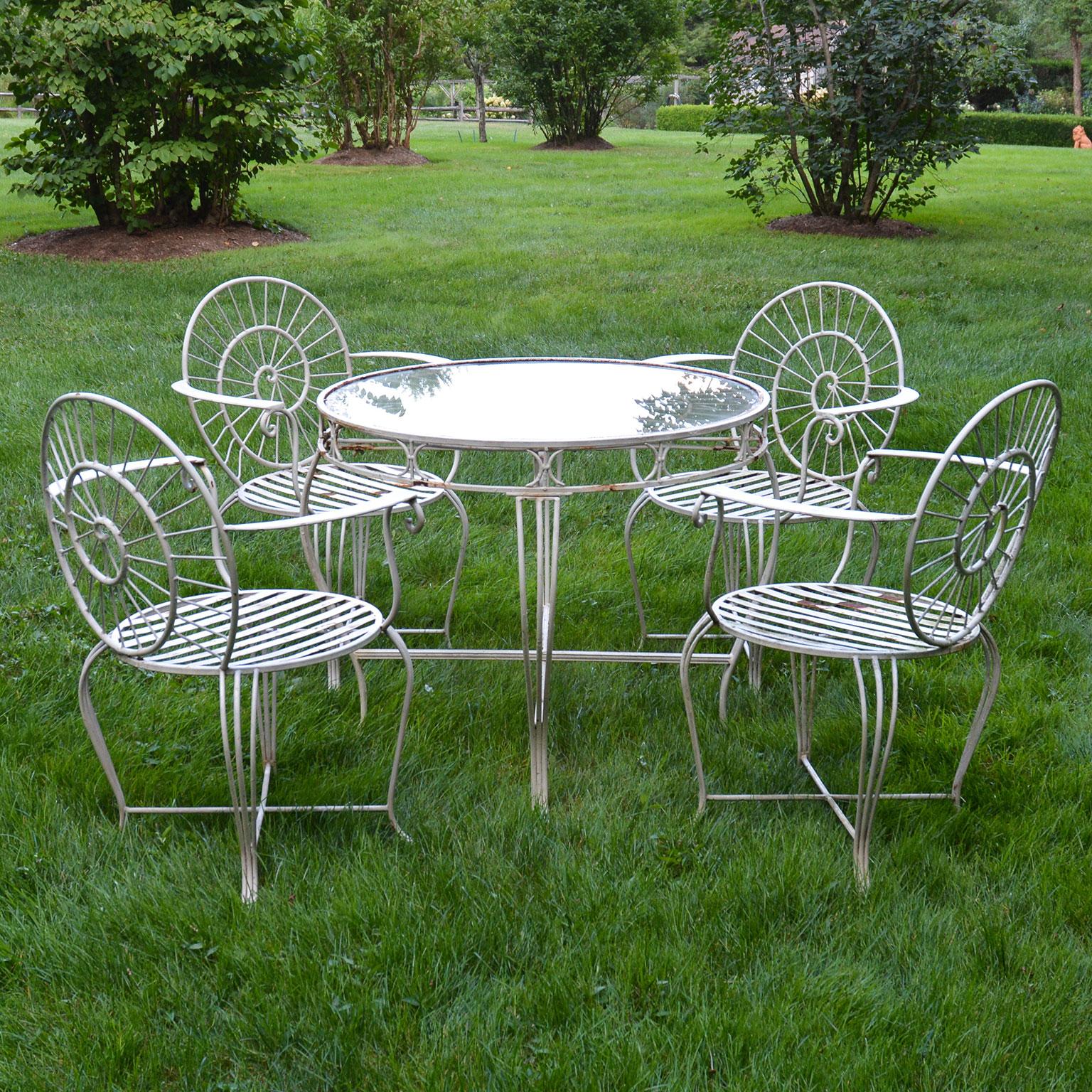 A wrought-iron dining suite comprised of four armchairs, the chairs with slatted seats, cabriole legs, and the chair backs with stylized nautilus shell design, the round table with cabriole legs and stylized nautilus top, with inset glass. Measures: