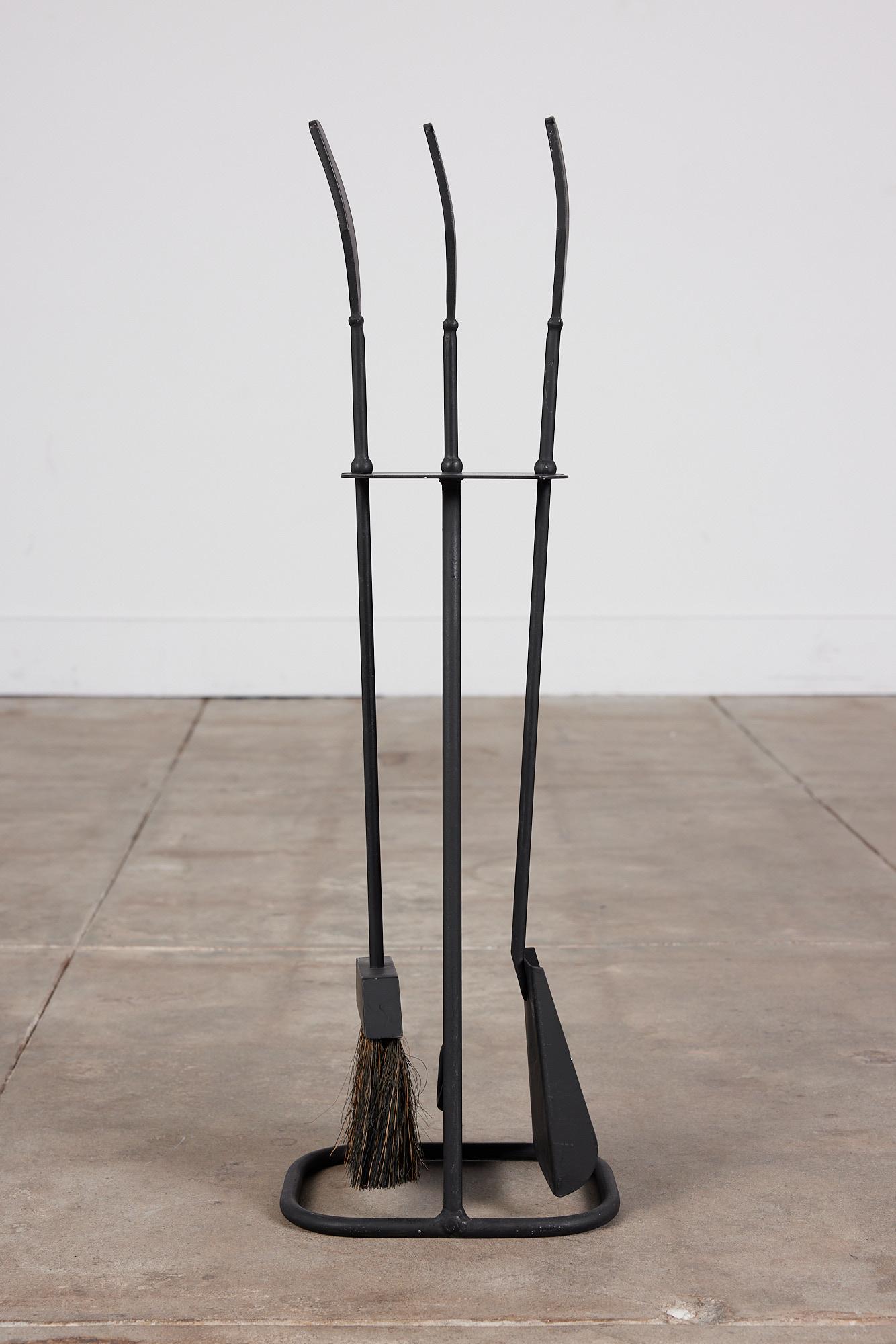 A three piece set of black wrought iron fireplace tools with stand in the style of Luther Conover. The set features three tools with flat curved handles and comes with a matching stand. All elements are made of blackened iron. This modernist piece