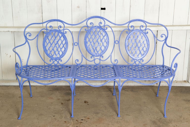 Charming baroque style wrought iron bench painted periwinkle. Lattice iron seat for three with three lattice ovals on the back. Eight legs, two curvy arms. Arm height: 26