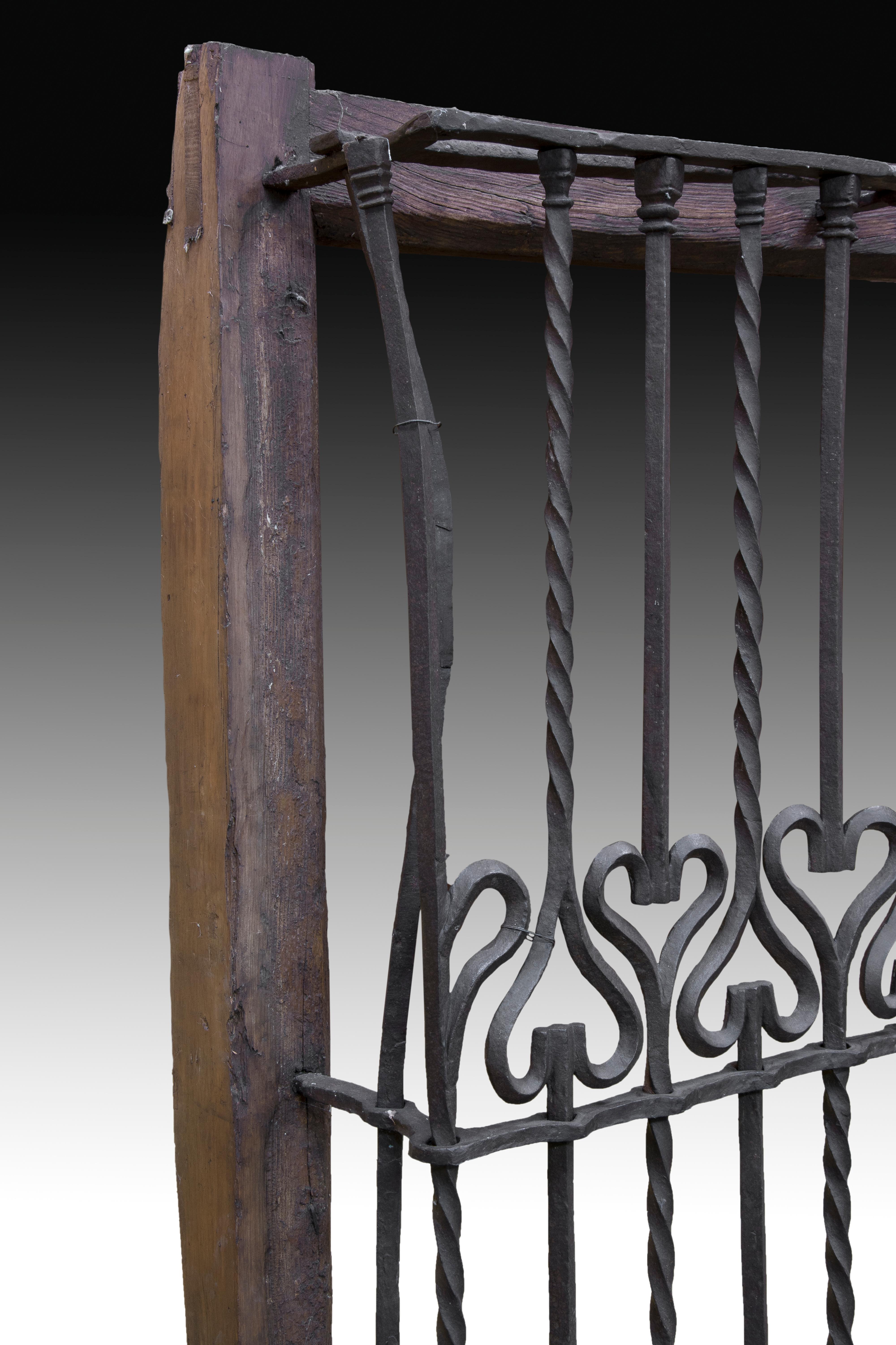 A carved wooden frame serves as a support for the wrought iron grille that has decorated balusters: all have a heart-shaped form in the centre, while the axes are smooth above and spiral below where the heart is upwards, and spirals up and smooth