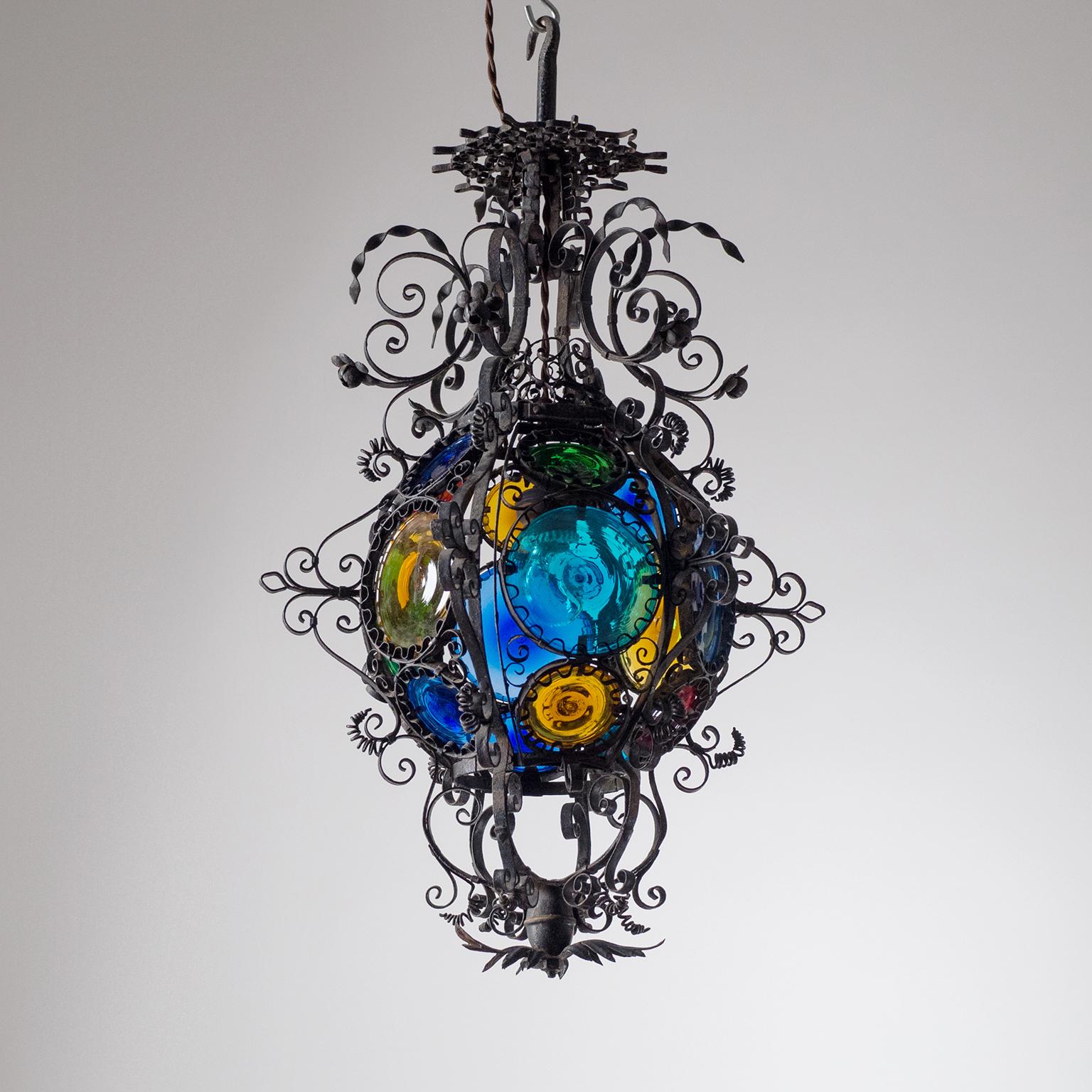 Lavishly decorated Italian wrought iron lantern with stained glass, late 19th century. A corpus (10inch/25cm diameter) of stained glass is surrounded by wrought iron curls, twists, floral and geometric ornaments. Nice original condition with patina