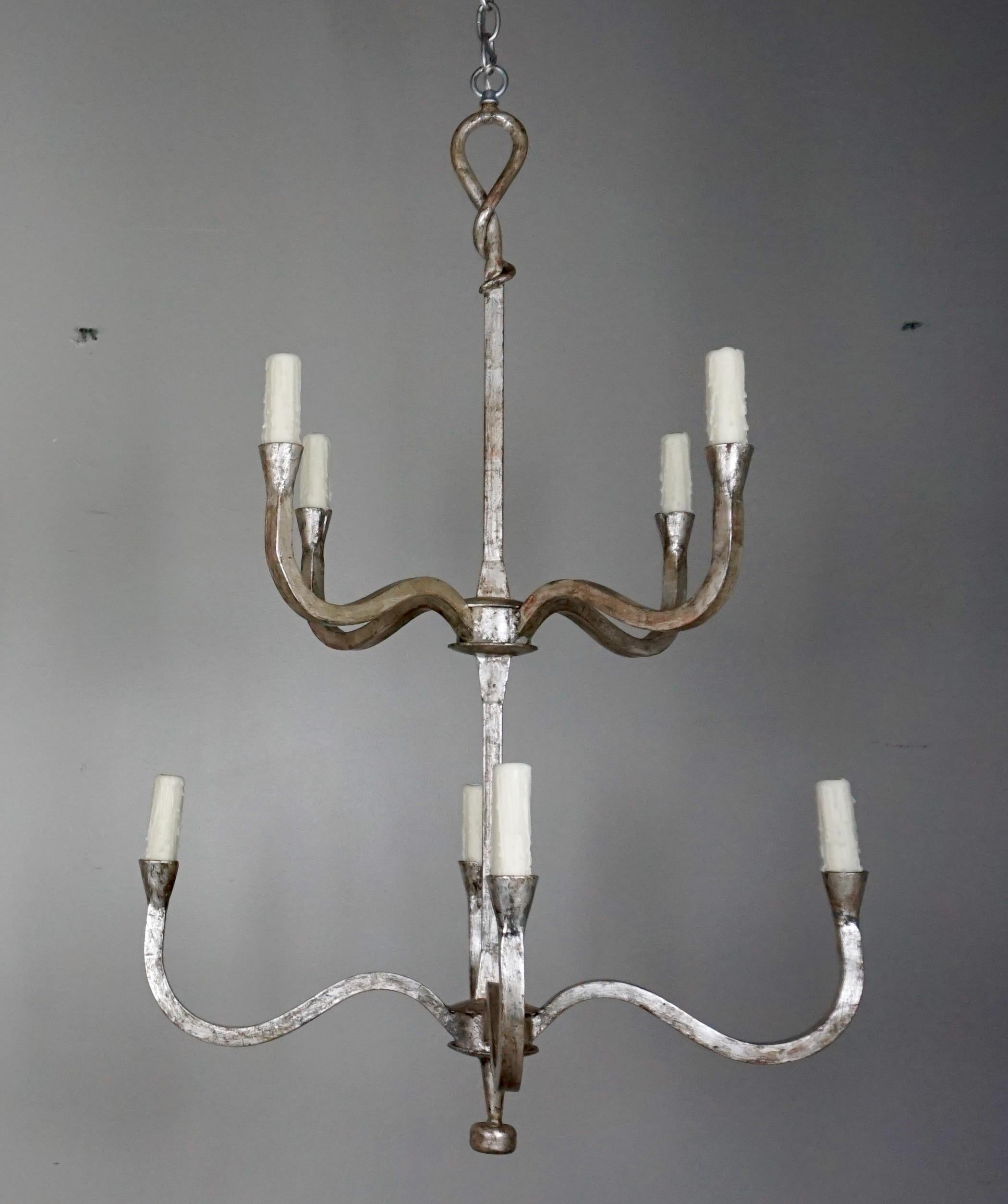 Pair of silver gilt wrought iron two-tier chandeliers. Each chandelier has eight candleholders with drip wax candle covers. The fixtures are wired and include chain and canopy. The fixture also comes in gold leaf a tete negre finish.