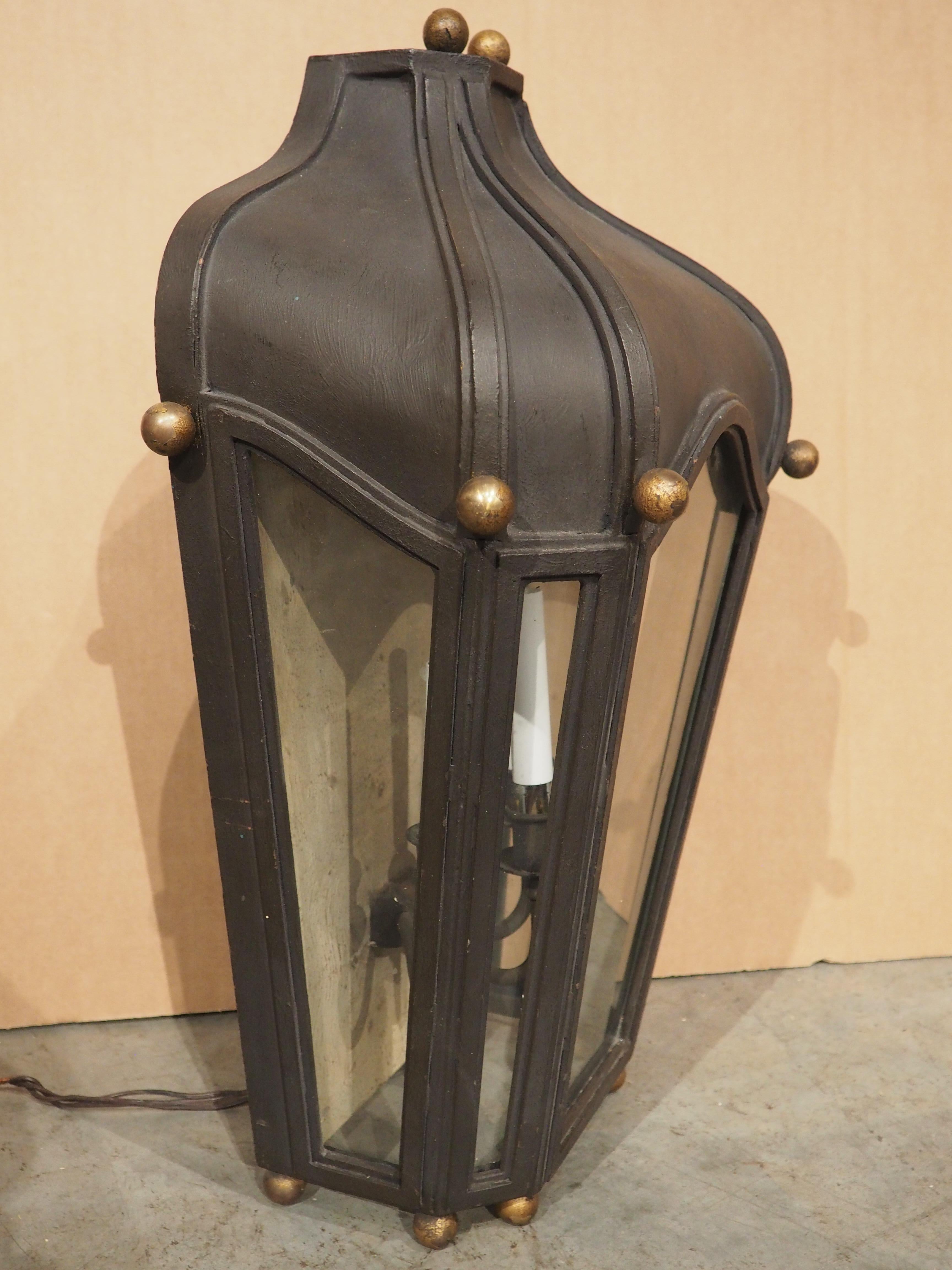 The flat back of this wrought iron lantern with glass allows it to be hung architecturally from any wall of the house. Five panes of glass enclose a three-arm sconce with saucer bobeches. Each arm has a gentle C-scroll shape beneath white faux
