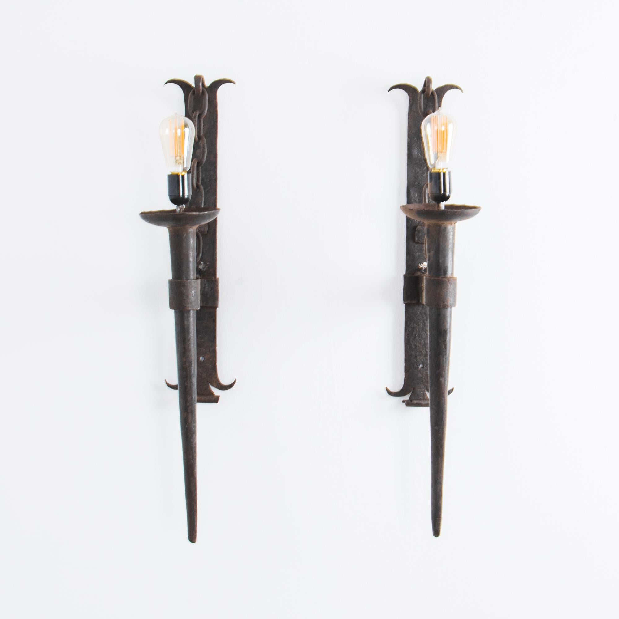 This pair of wall sconces from France, circa 1940, provide a striking light source. Made of wrought iron, their torch-like design gives a medieval gothic effect, accentuated by the chain detail on the wall brackets. Fully wired for use, these wall