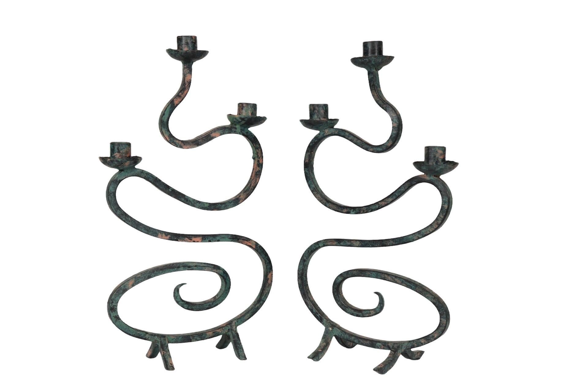 A pair of wrought iron wavy scrolled candelabra. Squared metal supports three bobeche and candle holders, undulating down into a loose scroll, terminating in a sharp point. Stands on splayed metal legs and painted in a verdigris green and copper