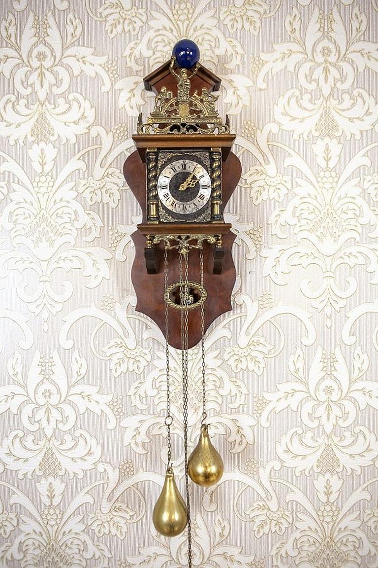 Wuba Wall Clock From the Mid-20th Century in Oak Case

The case is wooden, with glazed sides. Above the clock face, there is a brass crown with the maxim “Nu elck syn sin” which freely translates to “Now each to their own.”
The figurine of Atlas