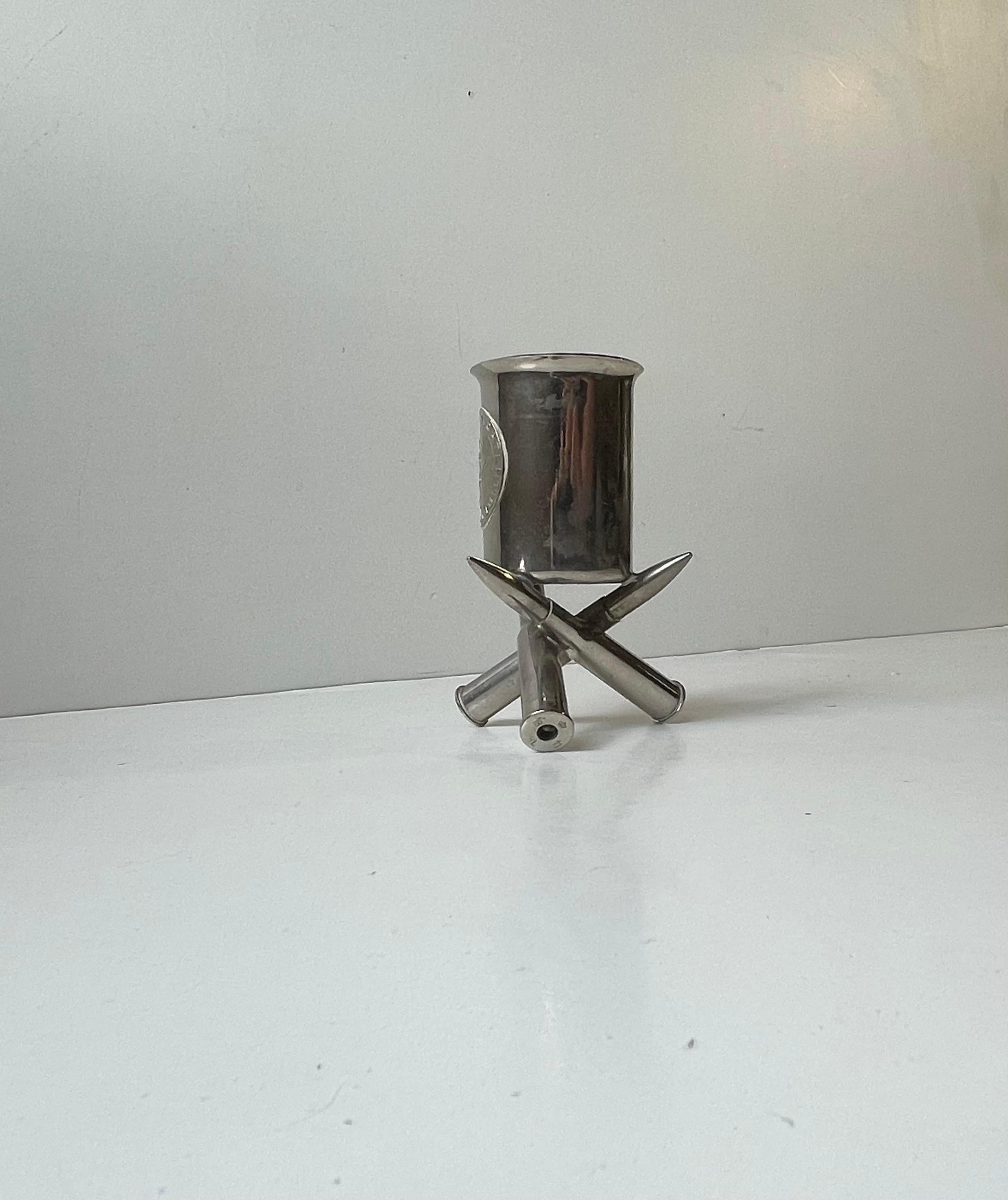 Un objeto del llamado arte de trinchera, con forma de portalápices o portacigarrillos plateado, fabricado con componentes de la época de la Primera Guerra Mundial. Las 3 balas de fogueo que componen sus patas tienen sellos del ejército danés y están